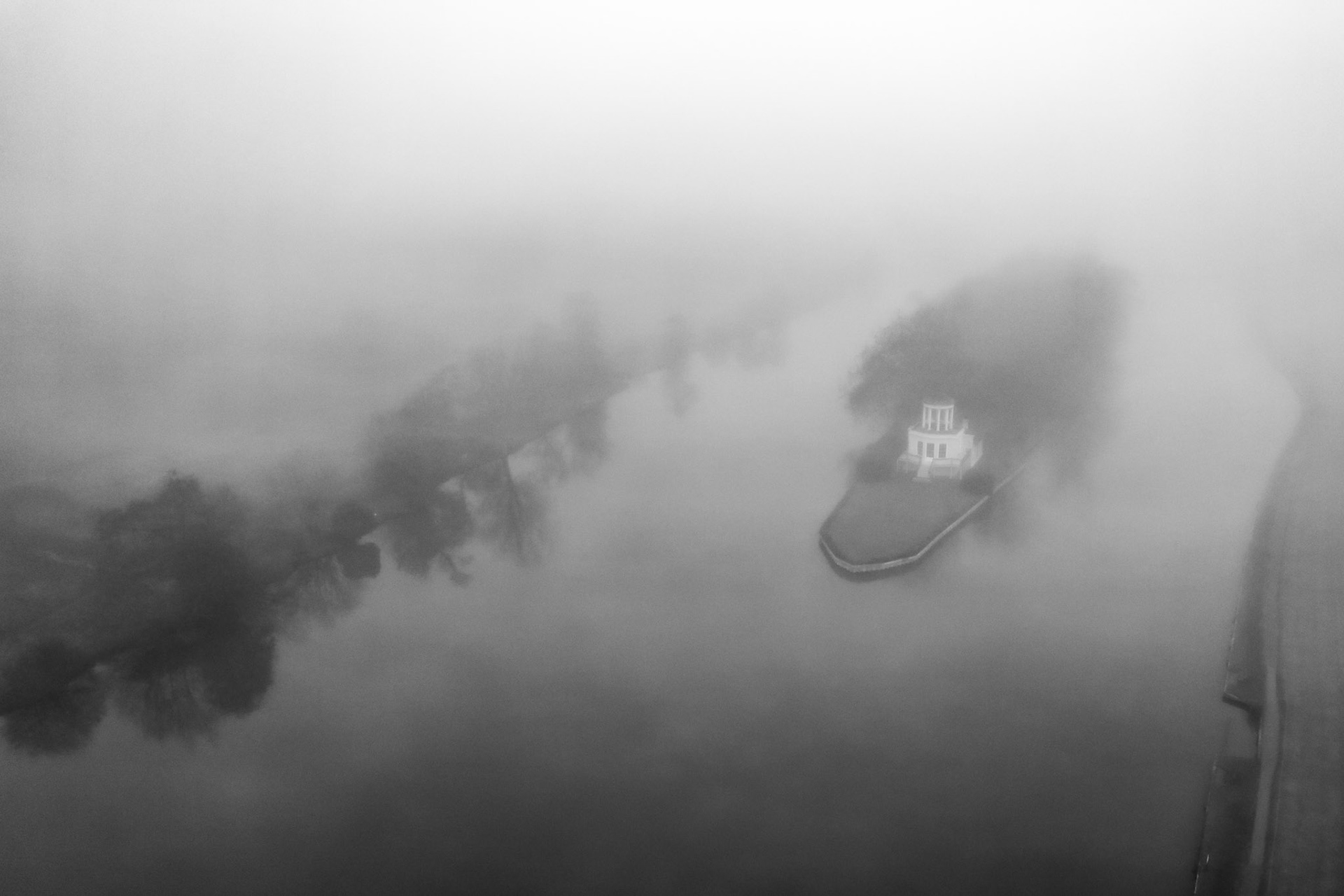 A dense fog inversion envelops the Thames at Henley, reducing a riverside property and its tree-lined peninsula to ghost forms suspended between obscurity and resolution. Shot from altitude at dawn in December, the monochrome conversion strips the scene to tone and shape alone, the lone white building acting as the sole anchor in an otherwise dissolved world.