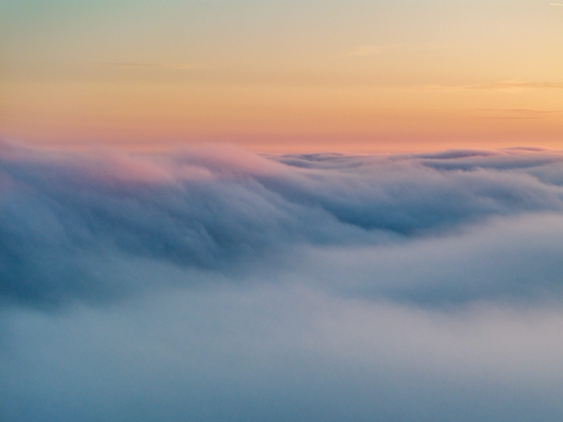 A temperature inversion buries the Thames Valley beneath a rolling sea of low cloud near Abingdon-on-Thames, the upper surface catching the last warm tones of a March sunset. Striations across the cloud top suggest gentle airflow, giving the otherwise featureless mass a quiet structural rhythm. The gradient from cool blue below to peach and teal at the horizon holds the eye without a conventional anchor point.