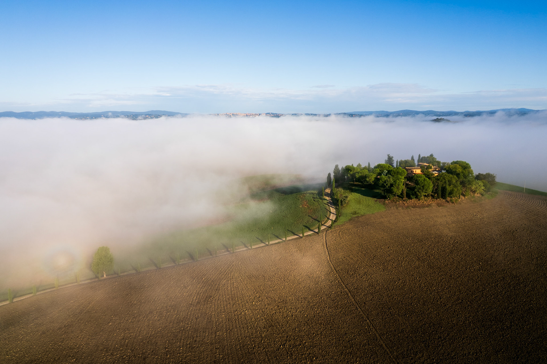 A Tuscan farmstead crowns a low hill above a valley fog inversion near Monteroni d'Arbia, Siena province. A cypress-lined track curves up from the mist-submerged plain, the ploughed field catching the warm early light while a distant hilltop town breaks the cloud horizon. The compression of earth, fog, and sky gives the composition an unusual stillness for an aerial frame.