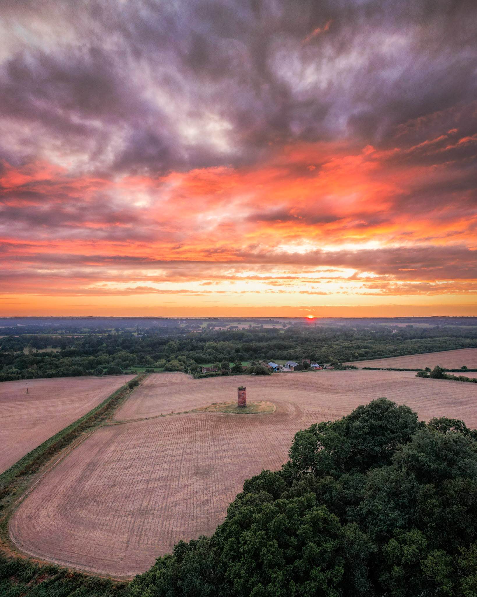 An isolated brick tower stands within a harvested arable field south of Reading, shot from altitude at the moment of sunset. Dramatic altocumulus cloud catches the last orange and violet light above a softly layered horizon of mixed farmland and woodland. The lone structure anchors an otherwise open composition, lending scale to the wide agricultural plain beneath.