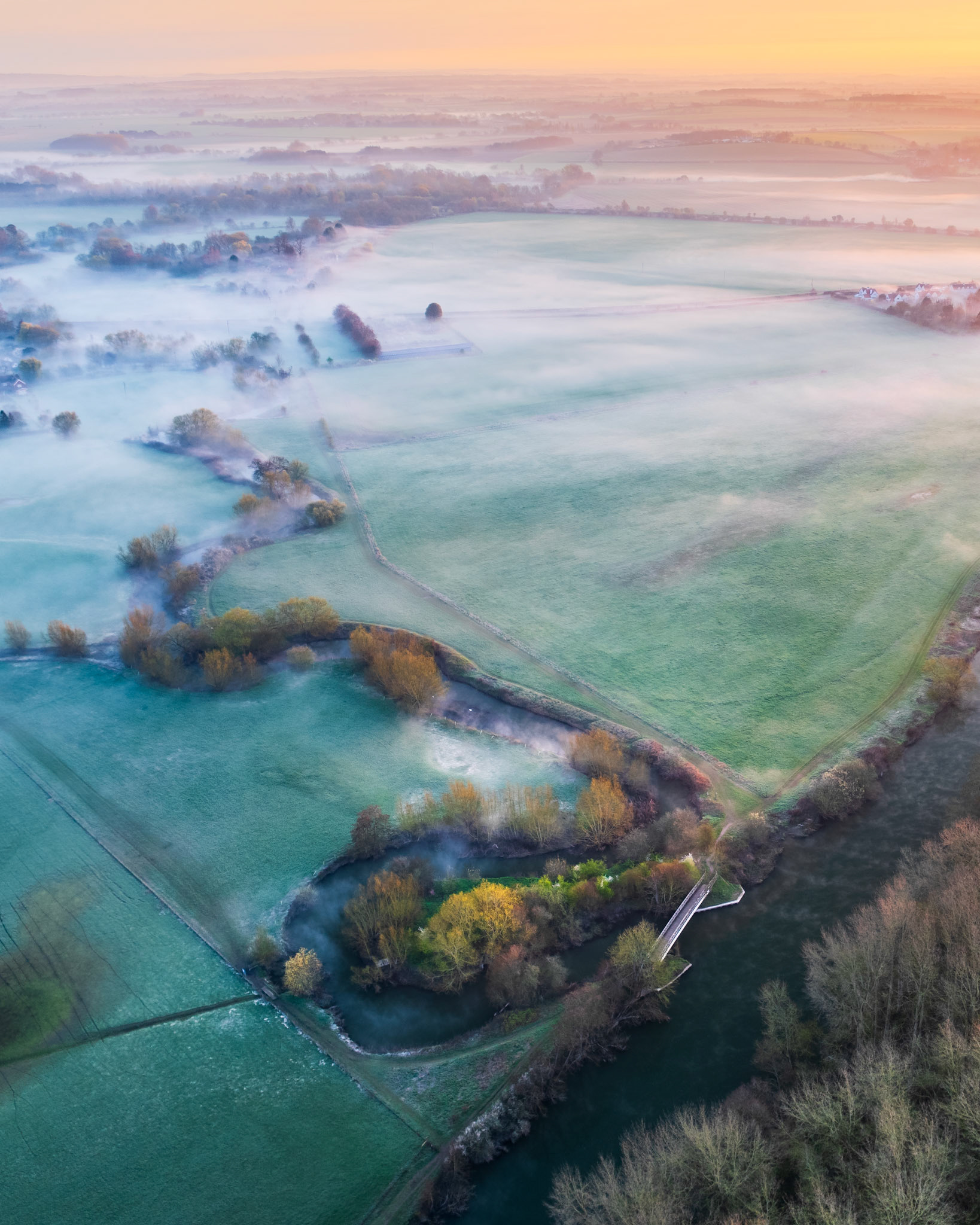 Ground fog pools across the Thames floodplain at dawn, seen from Brightwell Barrow near the Wittenham Clumps in Oxfordshire, the meandering river barely distinguishable beneath ribbons of mist that part around hedgerow trees and riverside vegetation. A small bridge marks a field boundary in the foreground, anchoring the geometry of pasture fields against the dissolving middle distance. The warm peach horizon above the fog layer completes an inversion of exceptional stillness.