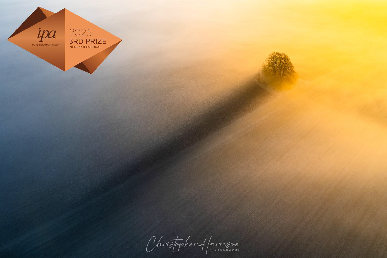 Christopher Harrison wins 3rd place in the Drone category of the International Photography Awards 2025 with his photograph "A Long Shadow". This image captures the moment the sunrise casts magnificent light through low lying mist and creates a beautiful shadow from a small clump of trees.
