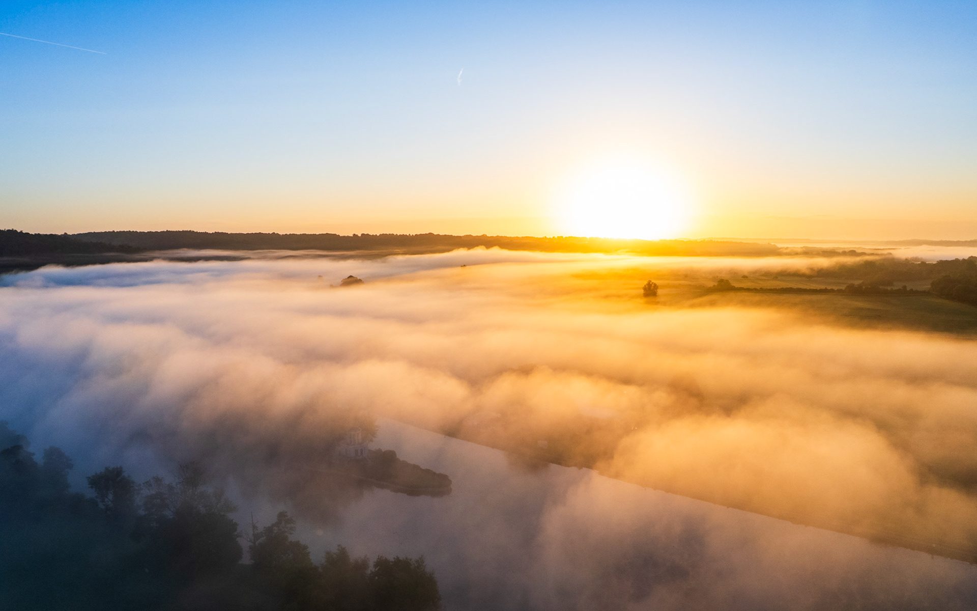 A temperature inversion fills the Thames Valley with dense ground fog at first light, tree canopies and hedgerows breaking the surface like islands. Photographed by drone near Henley-on-Thames in early autumn, the warm solar disc backlit the fog mass from the east, while cooler blue shadow held the foreground. The juxtaposition of luminous mist and dark emerging vegetation gives the scene an almost oceanic quality.