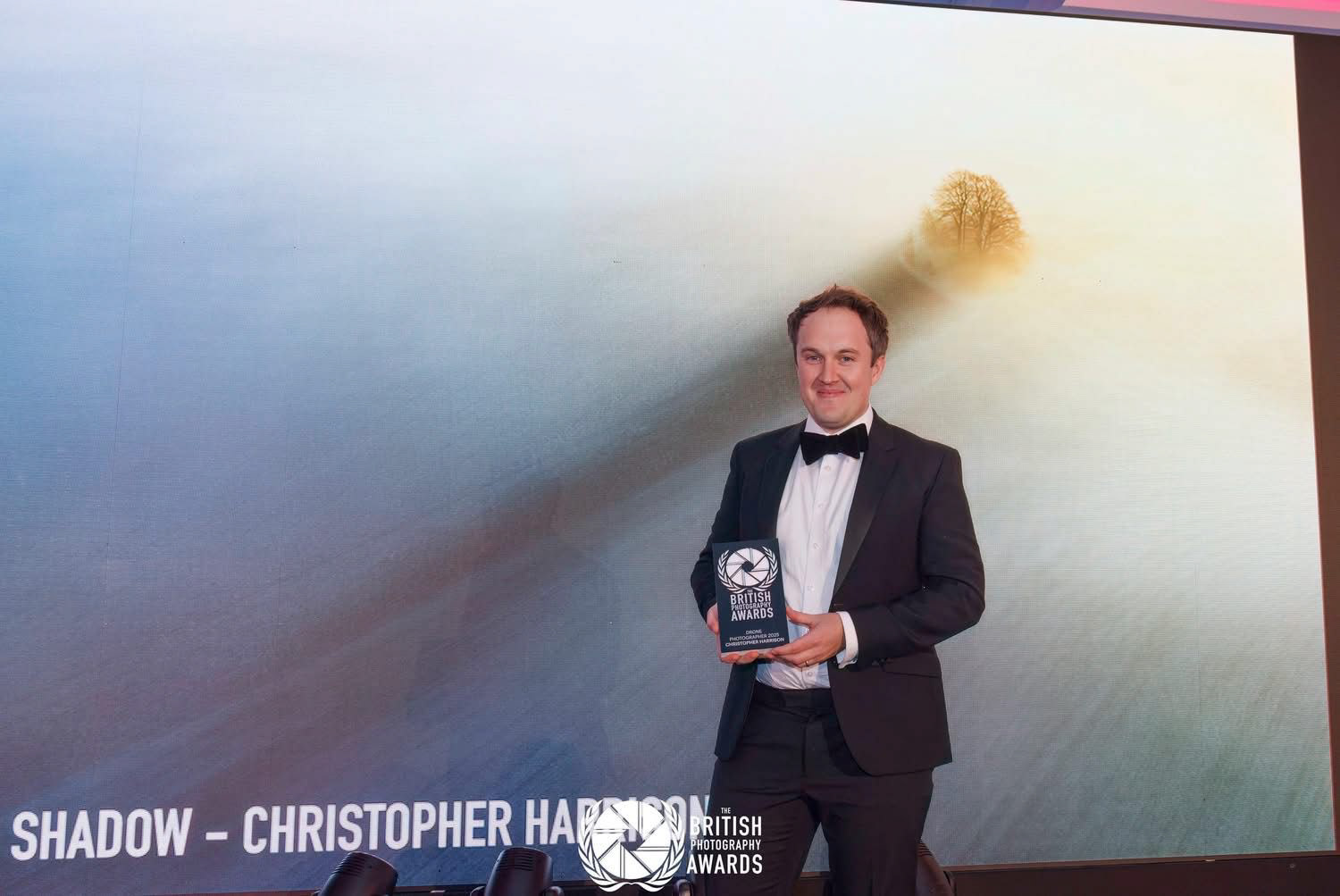 Christopher Harrison on stage at the British Photography Awards 2025, with his Drone Category Winner Trophy and his photograph The Long Shadow behind on the large screen.