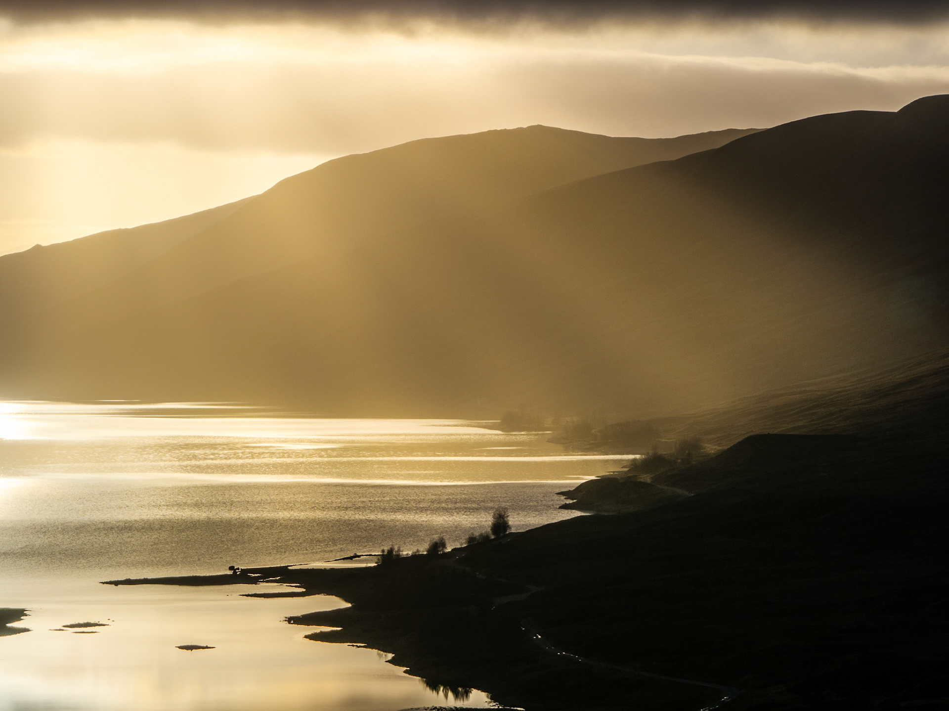 Crepuscular rays break through low cloud above a Scottish loch near Pitlochry, casting raking winter light across silhouetted hillsides and silver water. The aerial vantage compresses the shoreline into dark tonal bands against the luminous surface, while mist diffuses the far slopes into near-abstraction. Captured on Christmas Eve at low solar angle, the scene holds both scale and stillness.