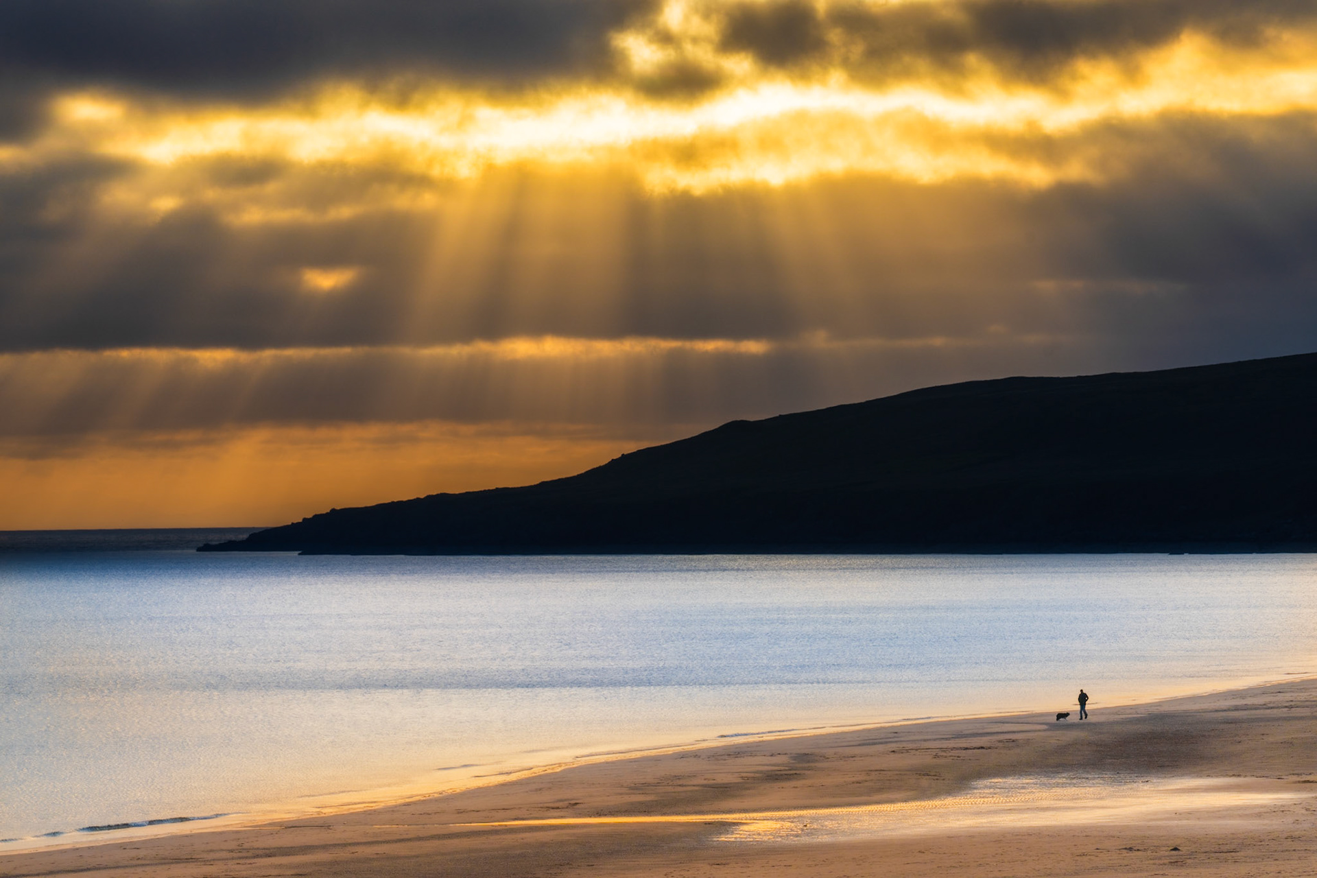 A man and dog take a walk on the stunning Big Sands Beach in rural North West Scotland, with epic beams shining through the clouds in the distance.