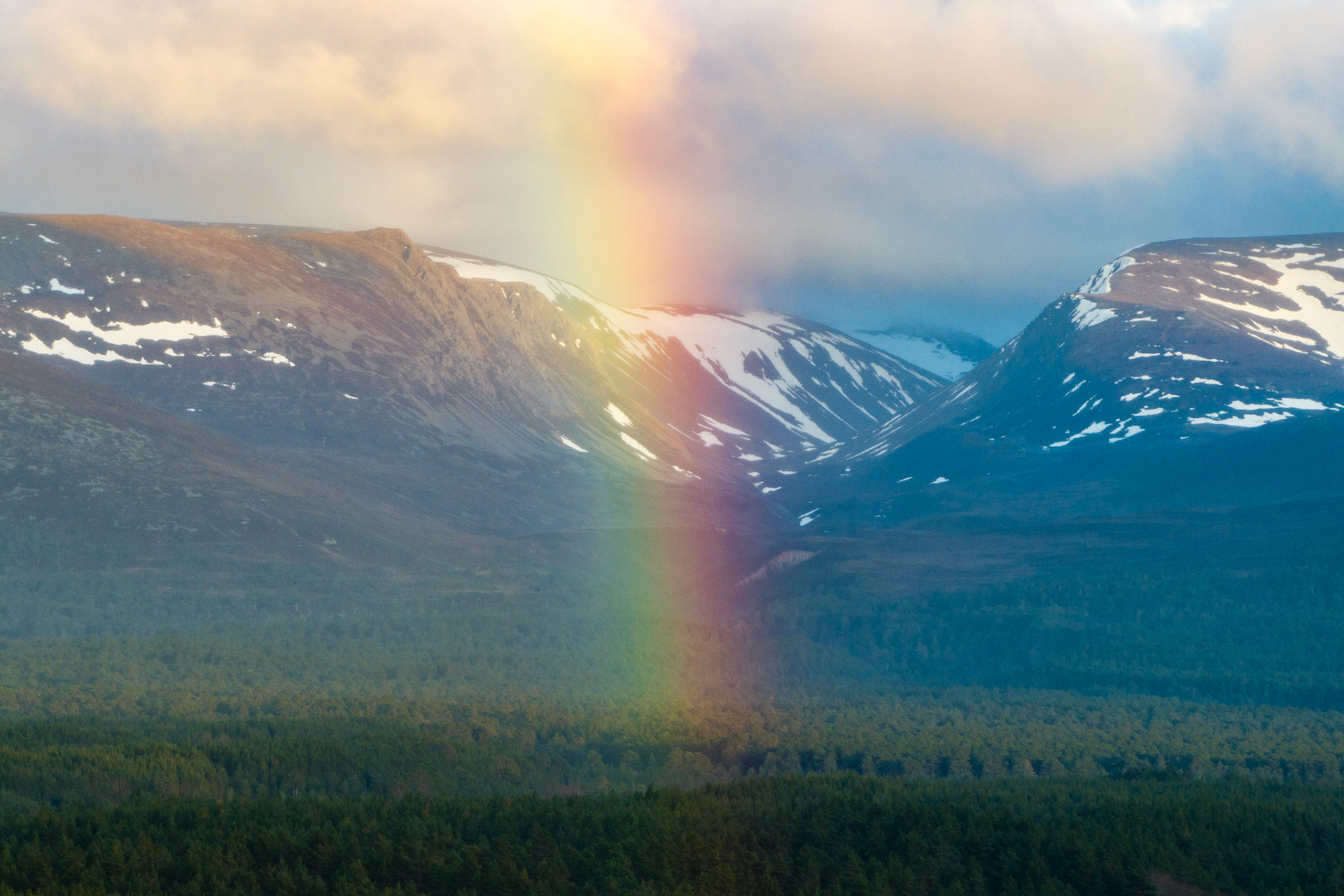 A partial rainbow descends into the mouth of a glacial pass in the Cairngorms, its arc anchored between snow-patched plateau flanks and the boreal pinewoods of upper Strathspey. The shaft of spectral light bisects the col with unusual precision, the surrounding cloud softening the plateau edges while spring snow persists on the higher ground. Captured from altitude in late evening light, the image exploits the elevated perspective to isolate the phenomenon within the mountain topography.