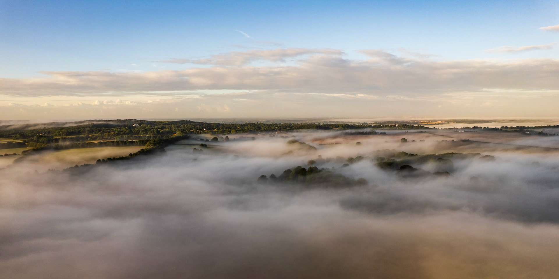 A temperature inversion pins a shallow fog across the Thames Valley lowlands near Maidenhead, submerging the fields while tree canopies and distant wooded ridges remain above the surface. Captured at first light in early September, the aerial vantage reveals the fog as a continuous, almost liquid plane, broken only by the crowns of mature broadleaf trees. The soft, diffuse illumination holds the scene in a quiet, pre-colour state.
