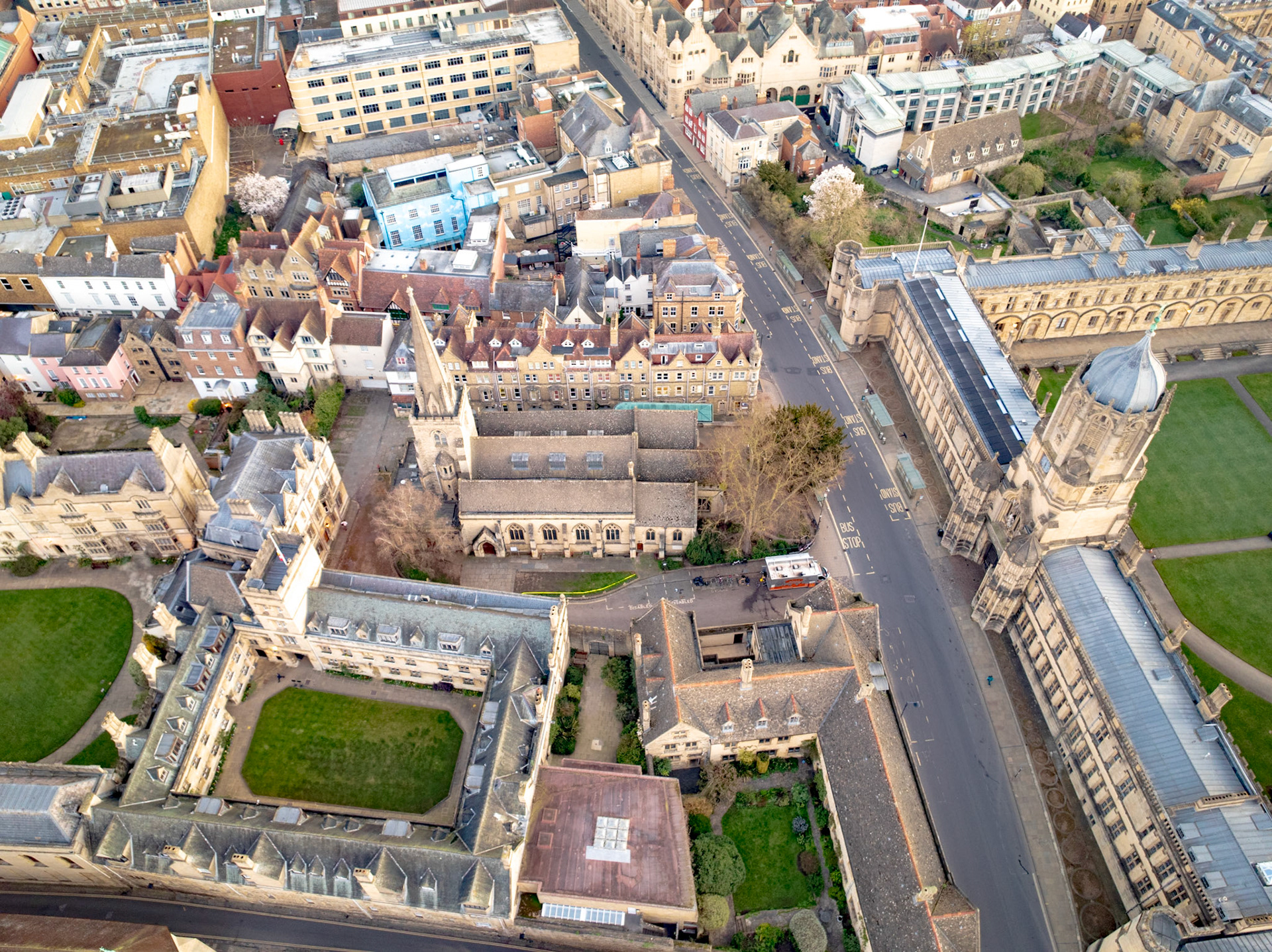 Oxford city centre seen from directly above in flat spring light, the quadrangles and rooflines of historic college buildings arranged in compressed, interlocking planes. The diagonal run of the High Street bisects the frame, anchoring the dense medieval and Georgian fabric against patches of college lawn. Flat overcast diffuses shadow and reduces tonal contrast across the scene.