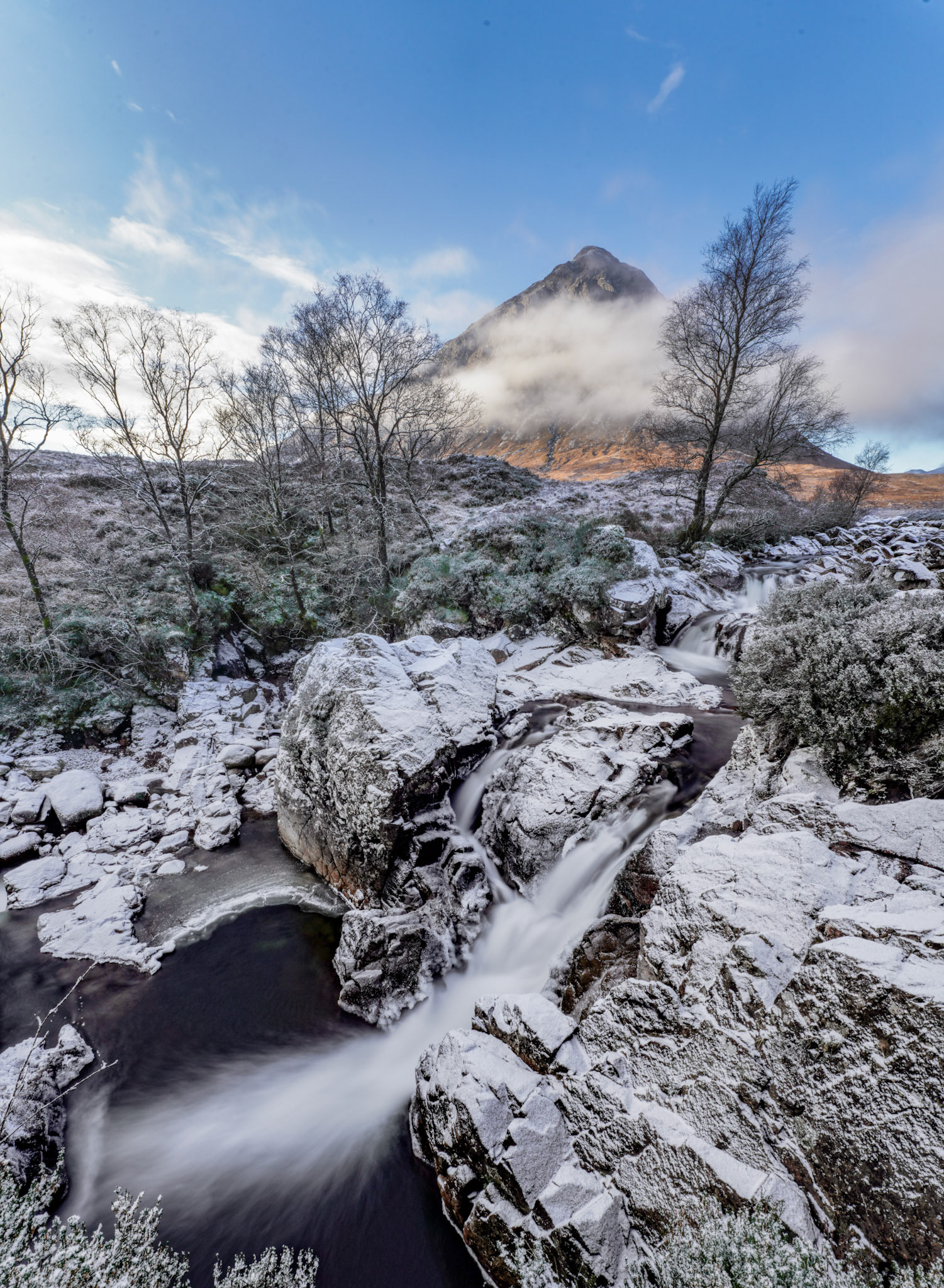 The River Coupall cascades over snow-dusted boulders below Buachaille Etive Mòr, its peak half-consumed by cloud. A long exposure renders the water as silk against the stillness of the frosted rockscape, the bare birches framing the pyramid summit with quiet insistence.