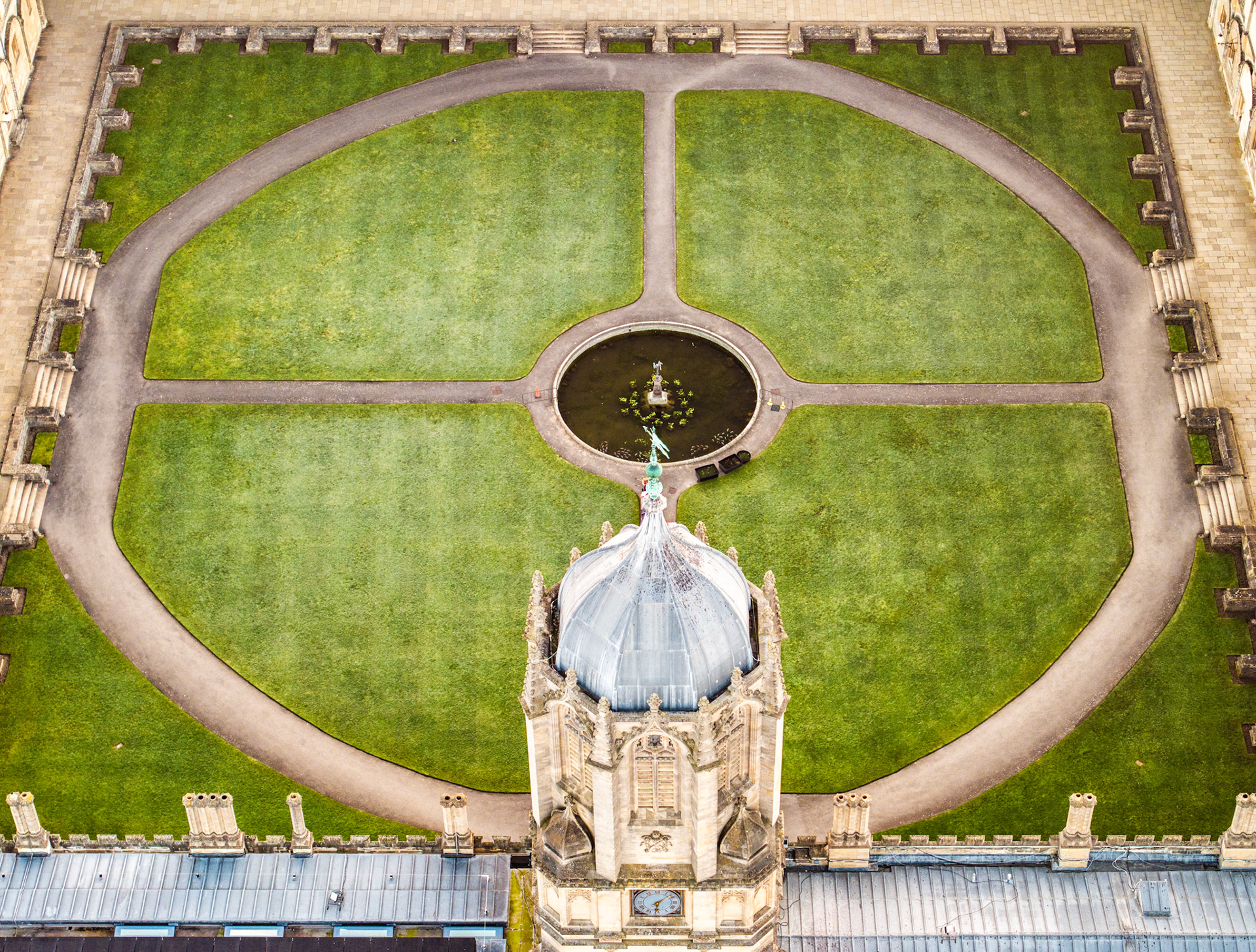 The dome of the Radcliffe Camera intrudes into the lower frame as a drone directly above traces the formal geometry of Radcliffe Square, Oxford. Circular gravel paths divide four quadrants of close-mown lawn, converging on a central fountain pool. The flat March light suppresses shadow and reduces the scene to near-diagrammatic form.