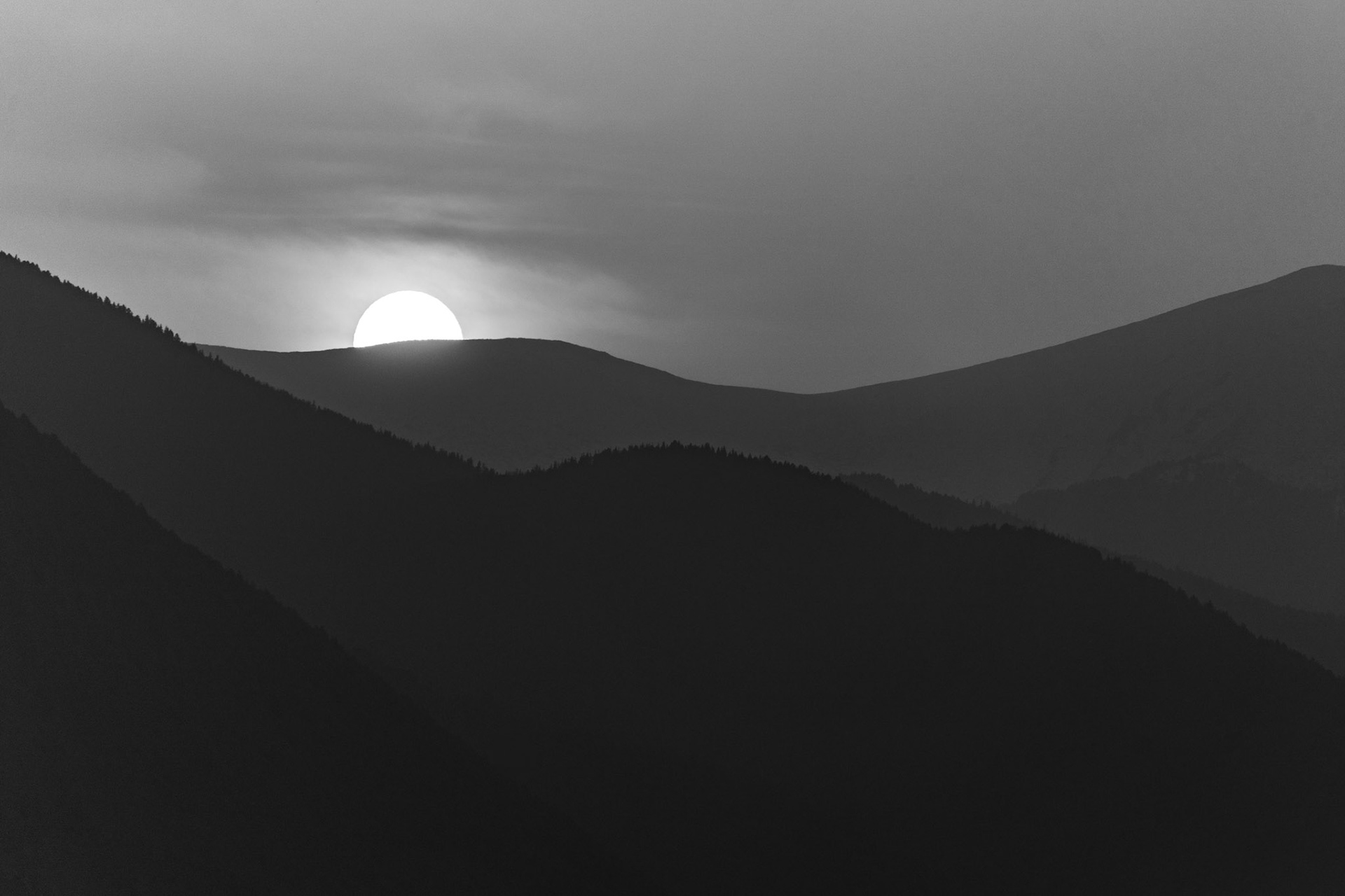 A low sun sits precisely on a moorland ridgeline, its disc rendered luminous against overcast cloud while successive hill silhouettes recede into atmospheric haze. The tonal layering — from near-black foreground slopes through mid-grey ridges to a bright corona — is handled with considerable restraint. Captured late afternoon in early spring, the image reads as monochrome conversion rather than in-camera black-and-white.