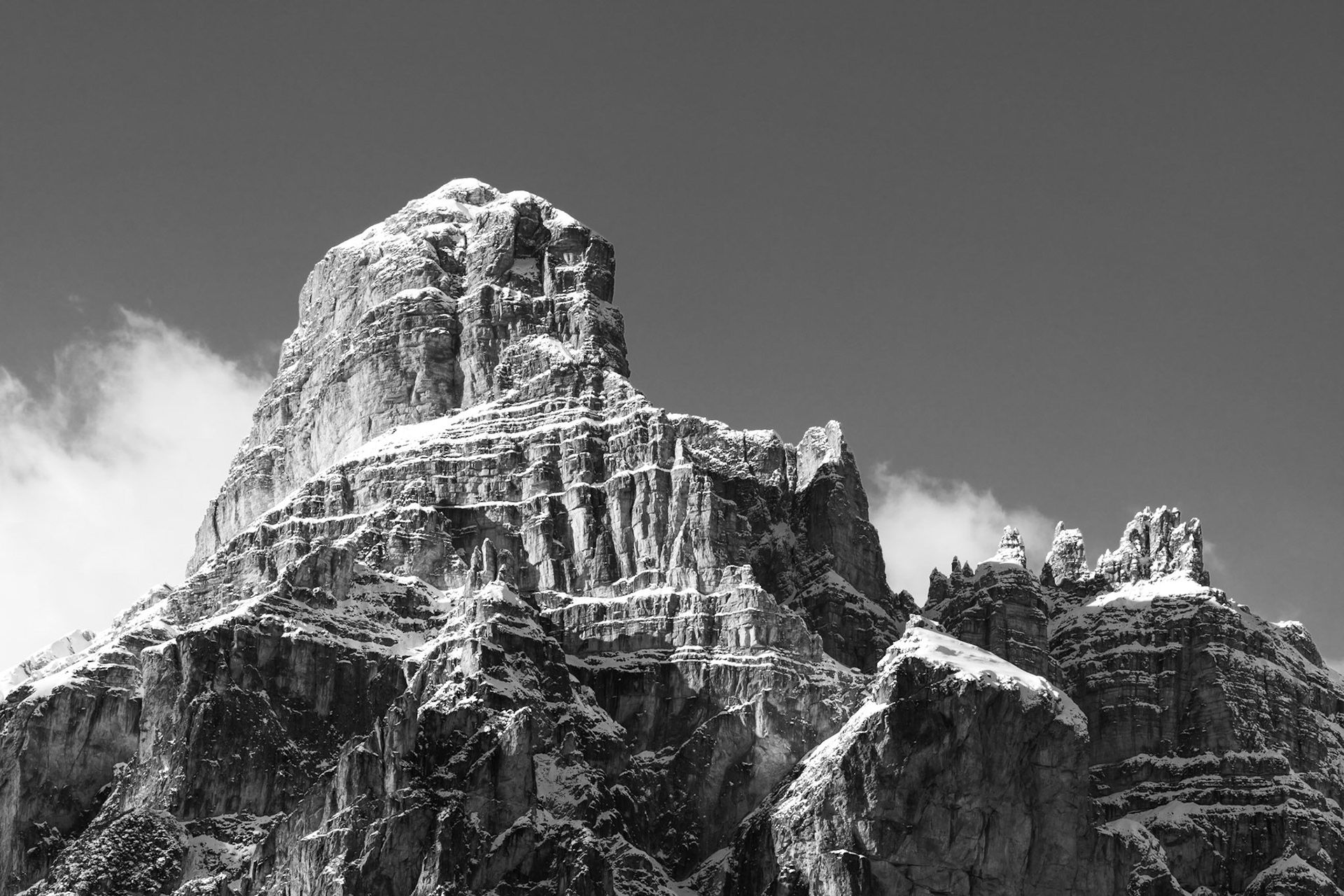 A monolithic dolomitic peak, heavily dusted with fresh snow, fills the frame against a dark cleared sky. The conversion to monochrome strips away colour distraction, allowing the stratified limestone geology and dramatic vertical relief to assert themselves fully. Wisps of cloud breaking around the secondary spires to the right suggest recent weather clearing.