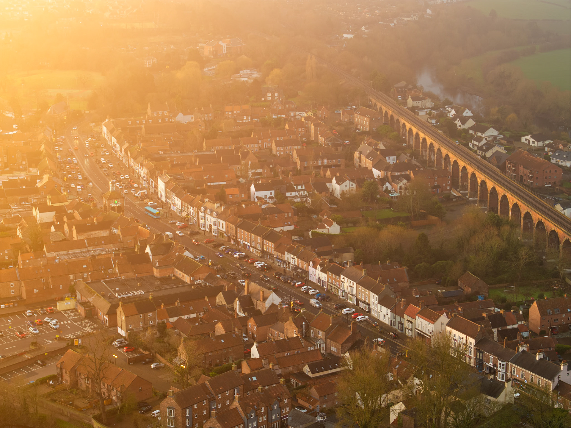 A Victorian railway viaduct arcs through a dense English market town, caught from altitude in the amber haze of a winter morning. The warm light unifies rooftops, high street, and arches into a single tonal plane, while mist softens the tree line to the north.