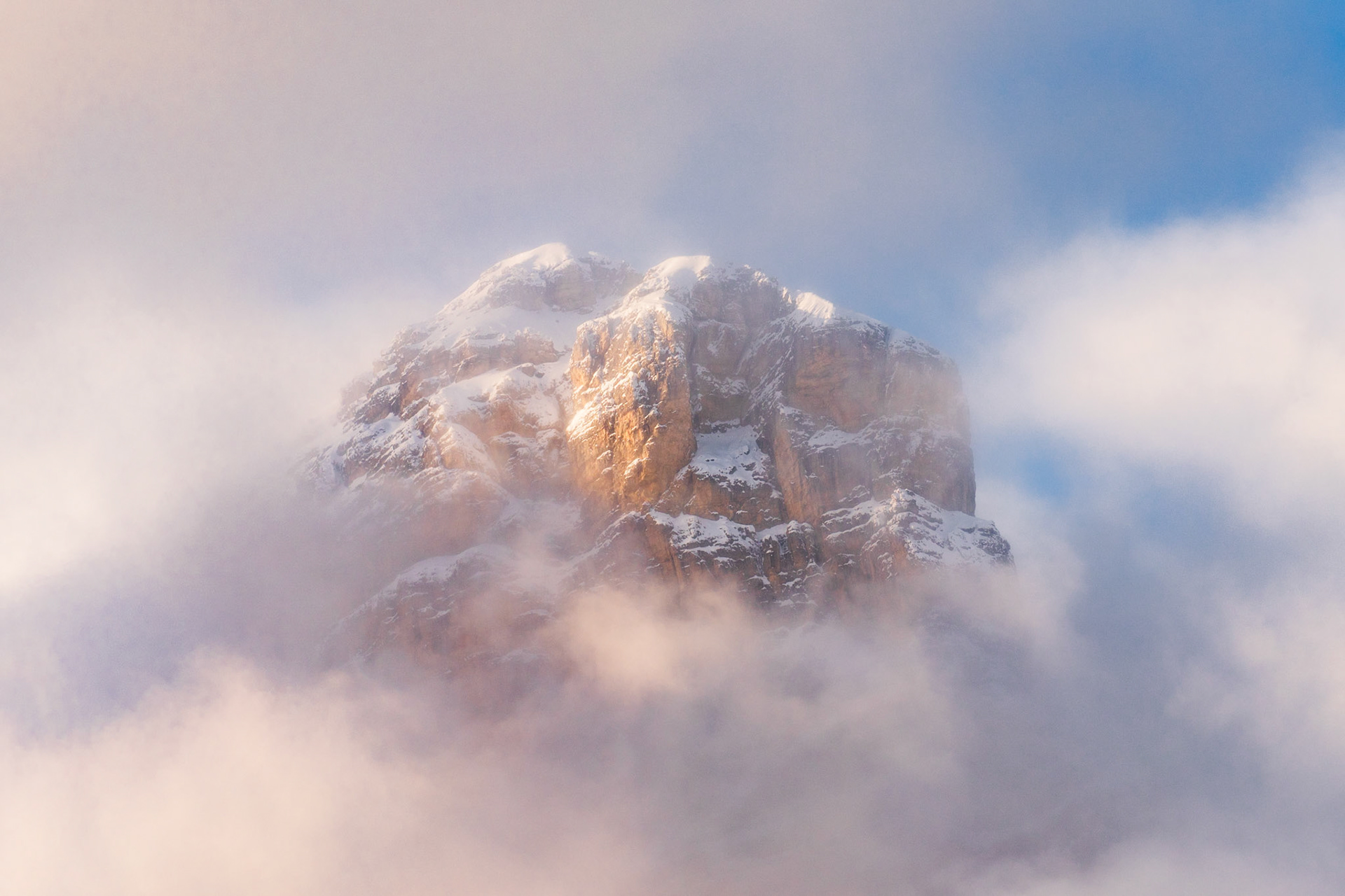 A snow-dusted rock summit breaks through a wrapping of low cloud, catching warm late-afternoon light on its vertical faces while the surrounding mist diffuses the scene to near-abstraction. The tight framing isolates the peak entirely from any skyline or foreground, lending it the quality of a floating mass. Captured in winter conditions, the contrast between warm ochre rock and cold white cloud carries considerable tonal tension.