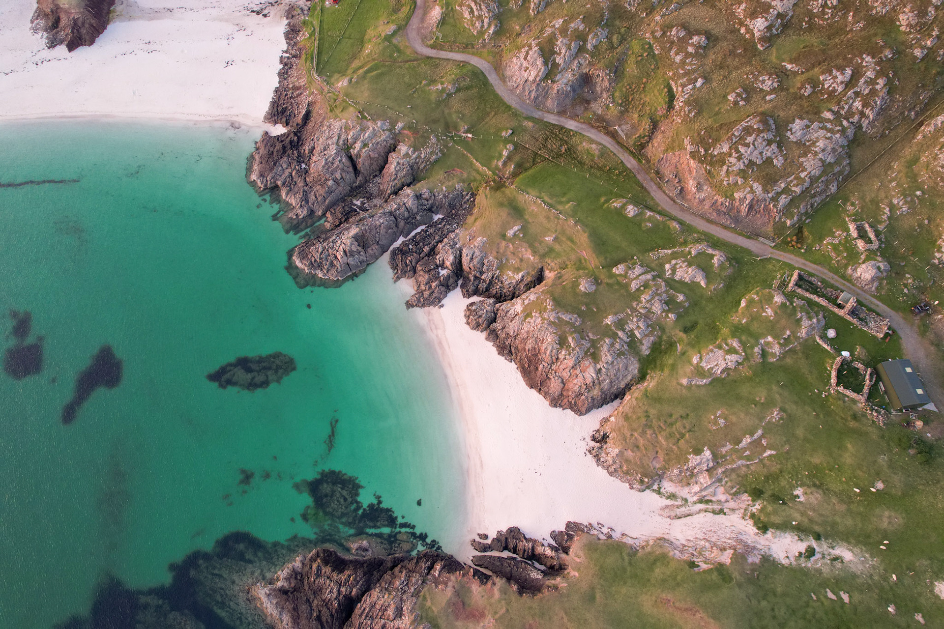 A small pocket beach of white shell sand lies sheltered between exposed gneiss headlands on the northwest Scottish coast, its turquoise shallows giving way to darker open water. Submerged kelp beds cast shadow patterns across the sea floor, while a single-track road and the remains of a stone building on the grassy hillside place this remote cove within a working, inhabited landscape. Shot vertically from altitude, the composition balances the geometry of rock, sand, and water without settling on a single dominant element.