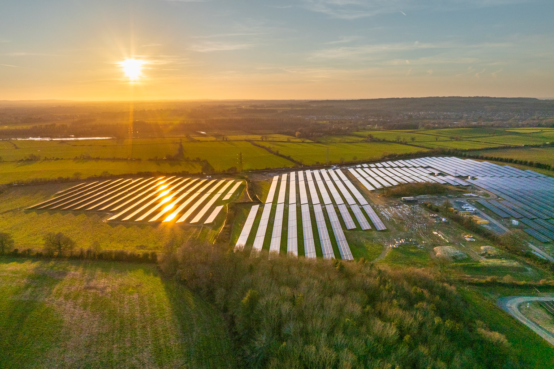 A large solar farm occupies former agricultural land on the Oxford floodplain, the low February sun catching the panel rows at a shallow angle and igniting a line of direct reflections across the left section. The surrounding patchwork of winter fields and hedgerows, with the Thames Valley settlements visible on the distant ridge, frames the geometry of the installation within a broader rural context.
