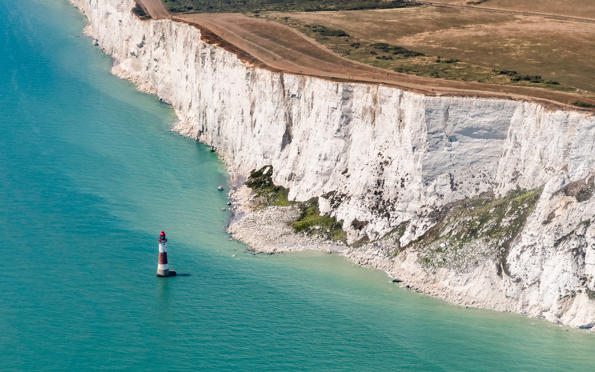 Beachy Head lighthouse stands isolated in calm English Channel waters below the sheer white chalk face of the Seven Sisters cliffs, East Sussex. The aerial perspective flattens the geometry of cliff, shore, and sea into near-abstract bands of colour, the lighthouse reduced to a small vertical accent against the turquoise. Midday light removes shadow relief from the cliff face, flattening texture in the brighter sections.