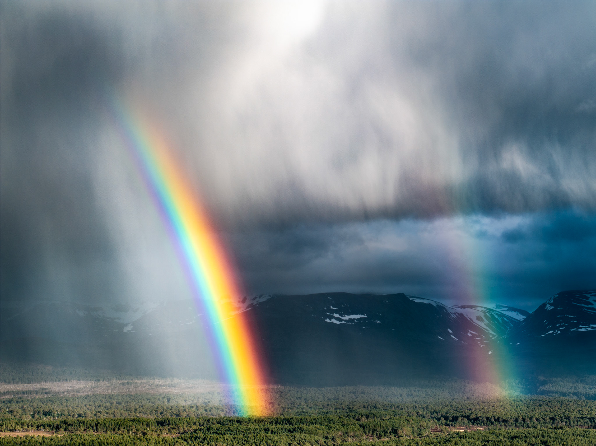 A vivid primary rainbow terminates above boreal forest in the Cairngorms, its arc set against the dark mass of a fast-moving convective storm. Snow-patched mountains anchor the middle distance while virga curtains the left frame, the aerial vantage isolating the optical phenomenon with unusual clarity.