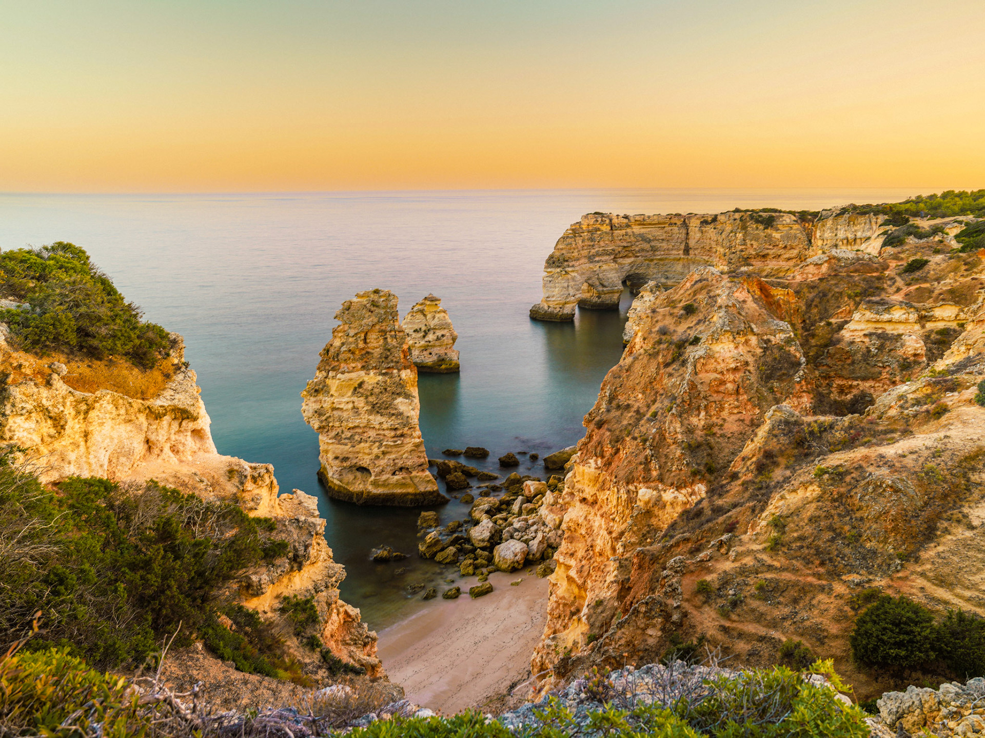 Twin sandstone stacks rise from a sheltered cove as the last light drains from a cloudless sky. The long exposure has smoothed the water to a pale mirror, emphasising the sculptural weight of the eroded cliffs that frame the scene. The location — characteristic golden limestone, layered cliffs, and sea-arch formations — is consistent with the Algarve coast of southern Portugal.