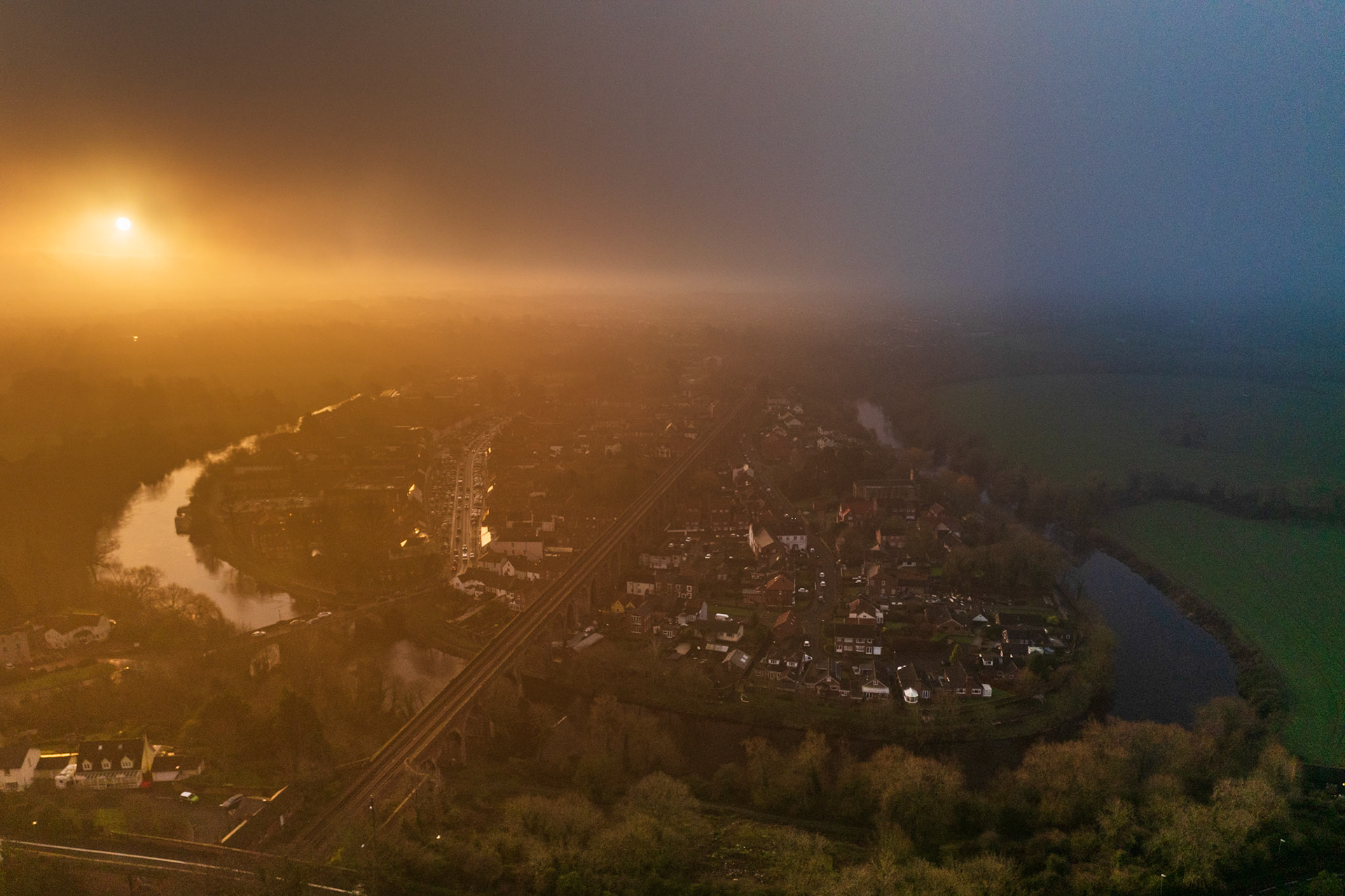 A temperature inversion splits the frame in two above a small English town encircled by a meandering river, the rising sun burning amber through fog to the west while cold blue shadow holds the eastern half. Captured from altitude in mid-winter, the hard colour boundary bisecting the scene gives the image its structural tension. Railway lines cut diagonally through the townscape below, grounding the aerial perspective.