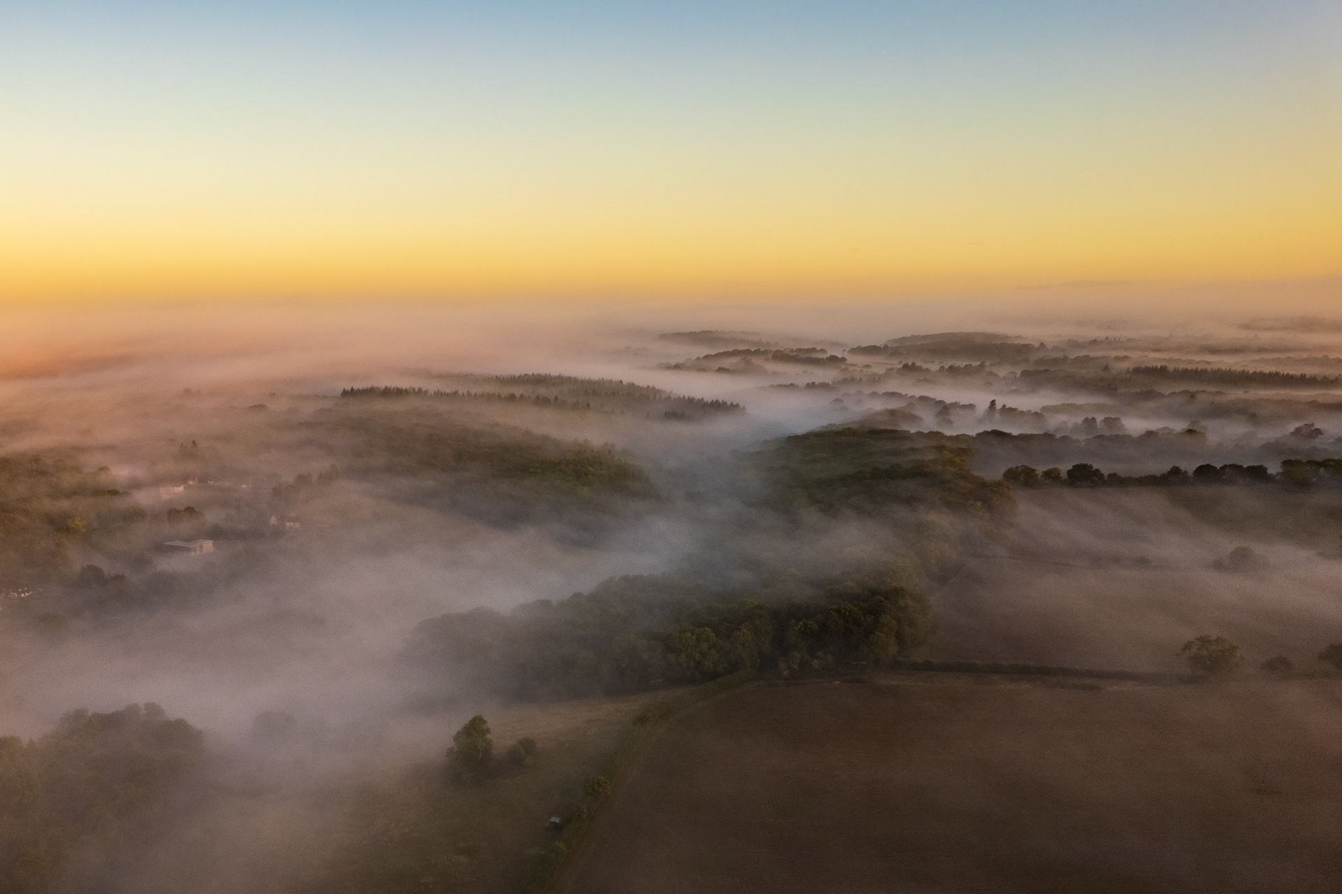 Ground mist pools in the valleys and hedgerow corridors of the Chiltern escarpment south of Henley-on-Thames, photographed from altitude at first light in early September. Tree canopies emerge as islands above the fog, their forms softened by the warm pre-sunrise glow. The layered recession of mist and ridge creates a quiet sense of depth across the rolling farmland.
