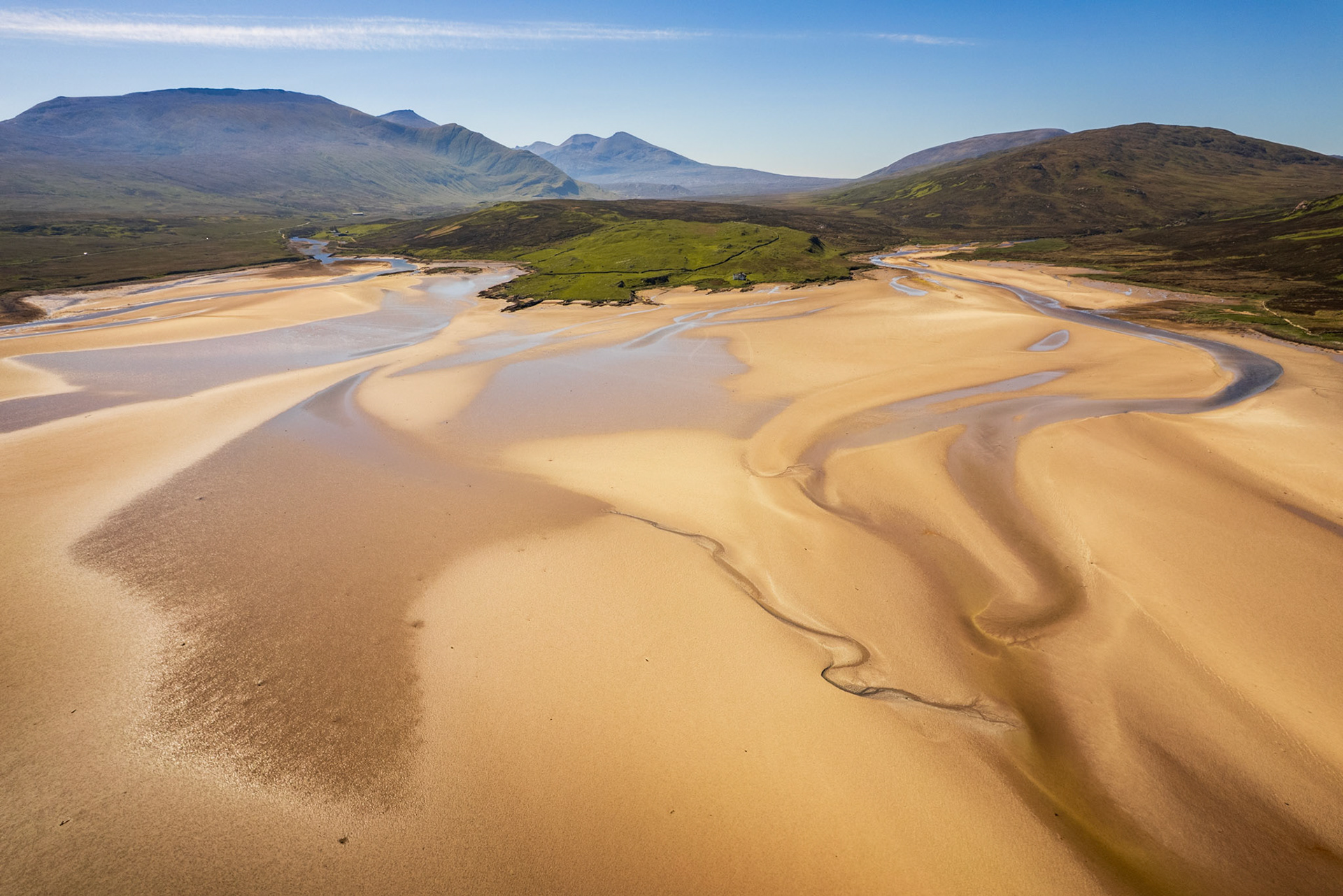 At low tide, braided channels carve sinuous lines across the tidal flats of a sea loch in Sutherland, the warm golden sand contrasting with thin slivers of pewter-grey water. Beyond, a patchwork of moorland and machair gives way to a receding line of mountains under a clear spring sky. The aerial vantage flattens the scene into near-abstraction while retaining the scale of the Highland interior.
