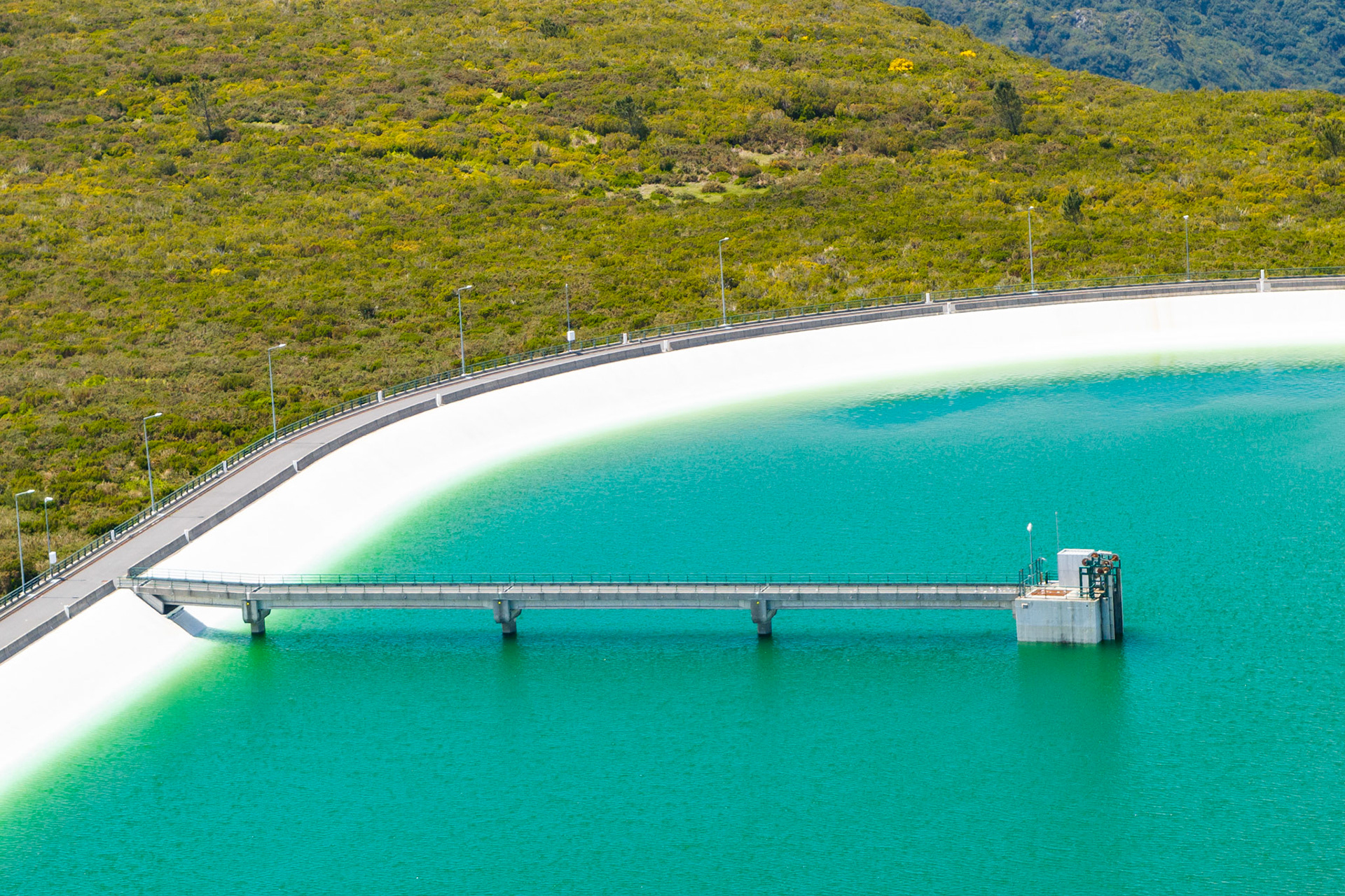 The curved face of the Calheta reservoir dam sweeps across the frame from an aerial vantage, its white concrete wall dividing vivid turquoise water from the scrubland hillside above. A narrow access bridge extends perpendicular to the intake tower, anchoring the geometric composition. Shot in bright overcast light that suppresses shadow and renders the water colour with unusual clarity.