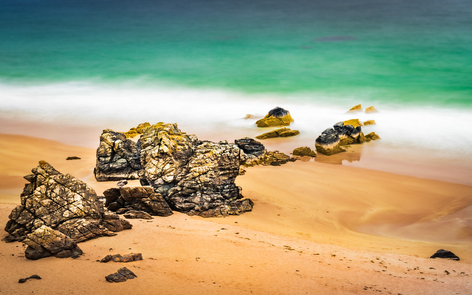 Fractured coastal rocks scatter across a sandy beach as long exposure reduces the incoming surf to a smooth, luminous wash. The vivid turquoise of the water contrasts with the warm ochre sand, the motion-blur lending an almost dreamlike stillness to an otherwise turbulent shoreline. Captured mid-afternoon in late spring.