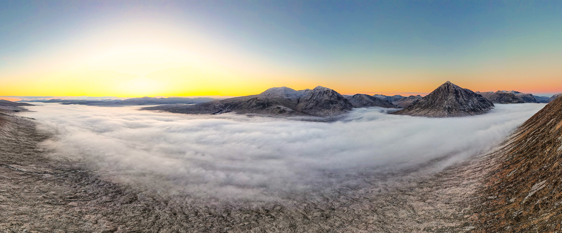 A temperature inversion fills a Highland glen to the brim with cloud, the summits of snow-dusted peaks emerging above the white floor at first light. The panoramic vantage from an elevated ridge compresses foreground frost-bleached moorland against a sky grading from amber to cold blue. The stillness of the inversion and the clean separation of layers give the scene an unusual geometric calm.