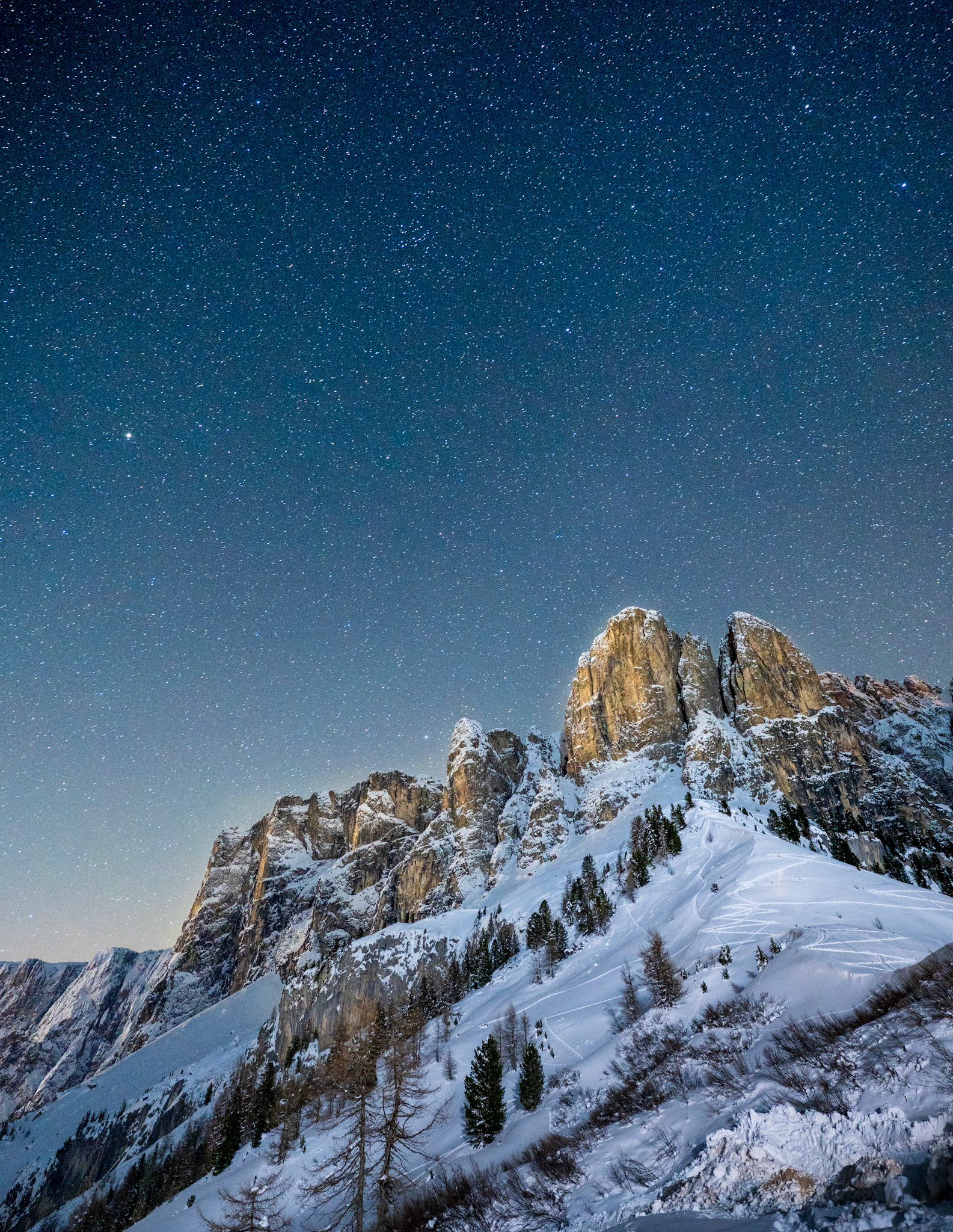 Moonlit dolomitic rock towers rise from winter snowfields beneath a densely starred sky, the warm amber of the illuminated cliff faces contrasting with the cold blue of the night. Ski tracks trace faint lines across the lower slopes, and scattered conifers punctuate the snow. The image balances terrestrial and celestial scale with quiet authority.