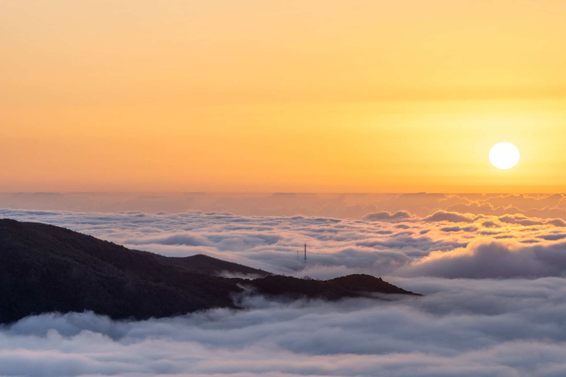 A temperature inversion fills the valleys below a mountain ridge, the cloud layer stretching unbroken to the horizon as the sun sits low and unobscured in a clear amber sky. A communications mast protrudes just above the cloud surface, a small note of the human world otherwise submerged. The image is taken from a high summit vantage at dusk or late evening in late May.
