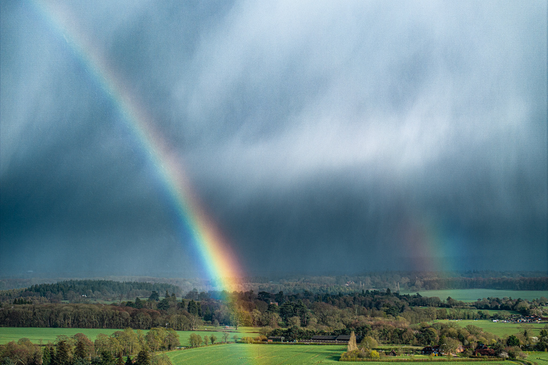A vivid primary rainbow descends through the base of a storm cell above the Thames Valley near Henley, its foot illuminated by a shaft of low evening light breaking beneath the advancing cloud. The aerial vantage compresses the arc against a dark curtain of rain, while spring fields and deciduous woodland spread beneath in fresh growth. The convergence of storm shadow and spectral colour gives the image an uncommon intensity.