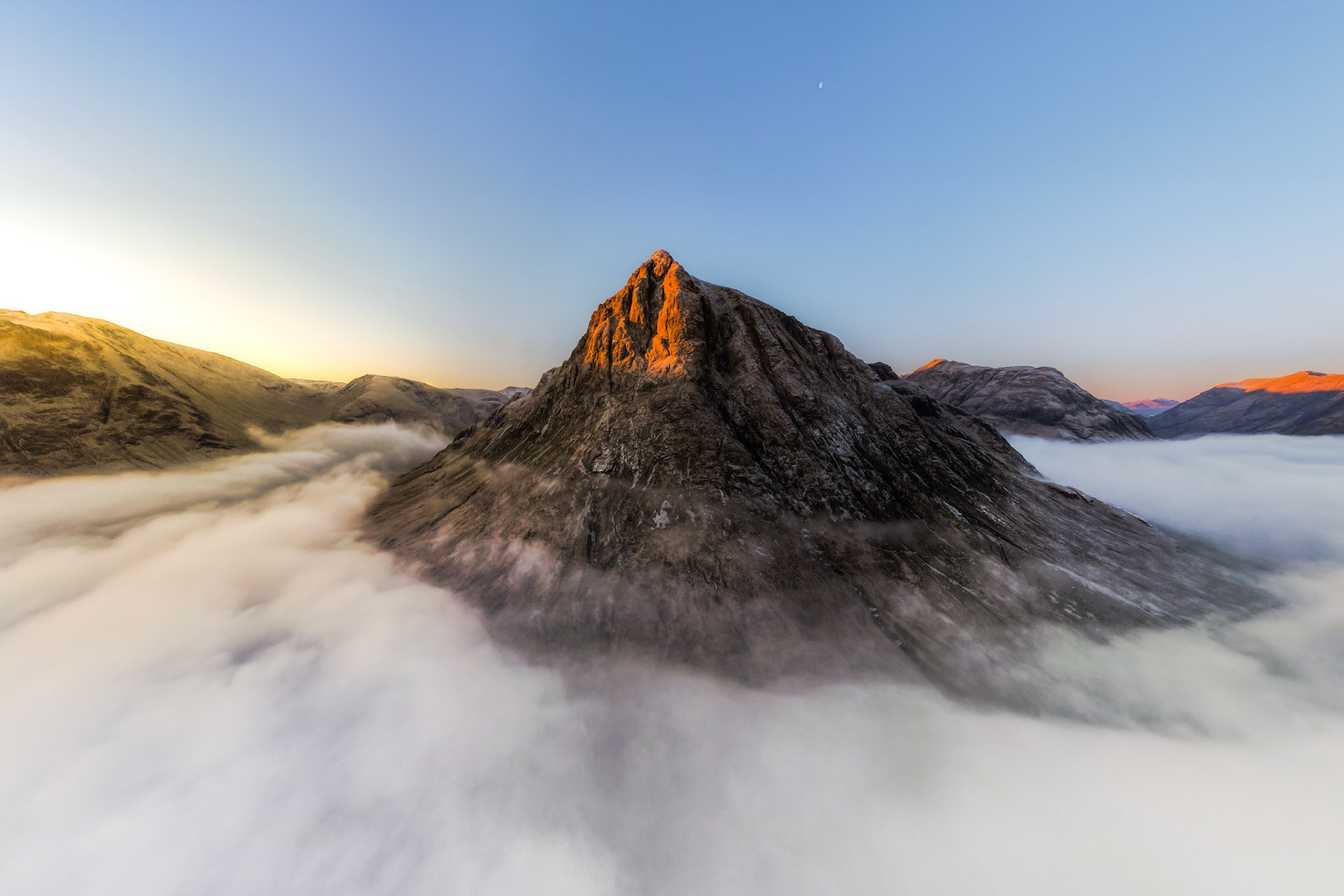 A temperature inversion buries the glens beneath a still sea of cloud, leaving the rocky summit of Sgorr Dhonuill emerging into first light above Ballachulish, Scottish Highlands. The crescent moon holds its position in a steel-blue sky as alpenglow catches the peak's upper ridgeline. The aerial vantage compresses the surrounding mountains into a silent archipelago of stone.