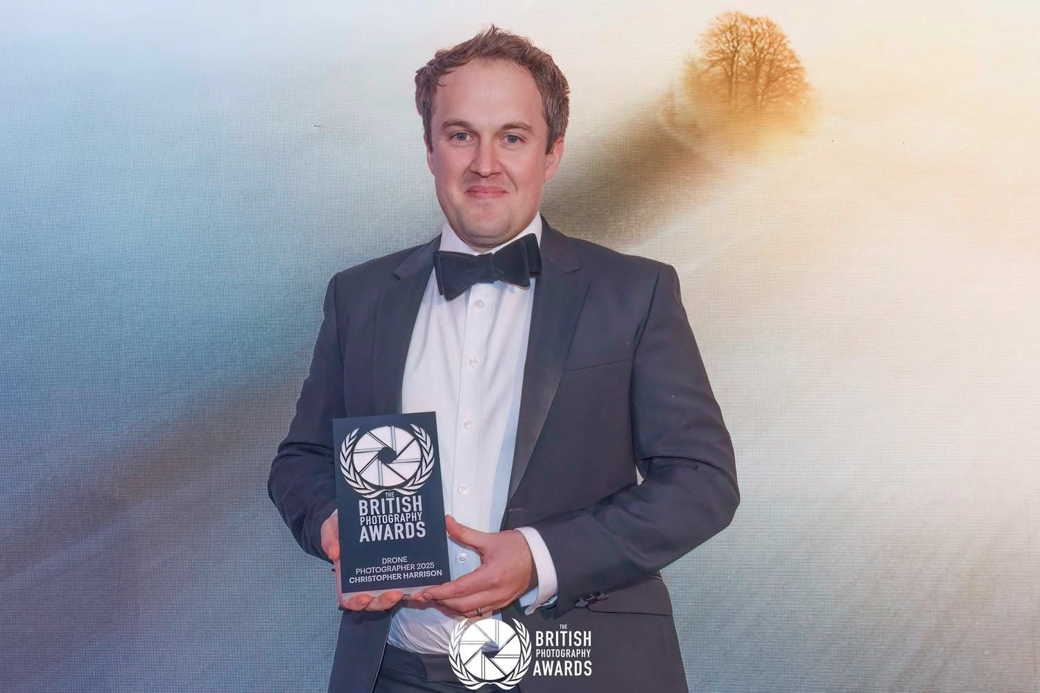 Christopher Harrison on stage at the British Photography Awards 2025, with his Drone Category Winner Trophy and his photograph The Long Shadow behind on the large screen.
