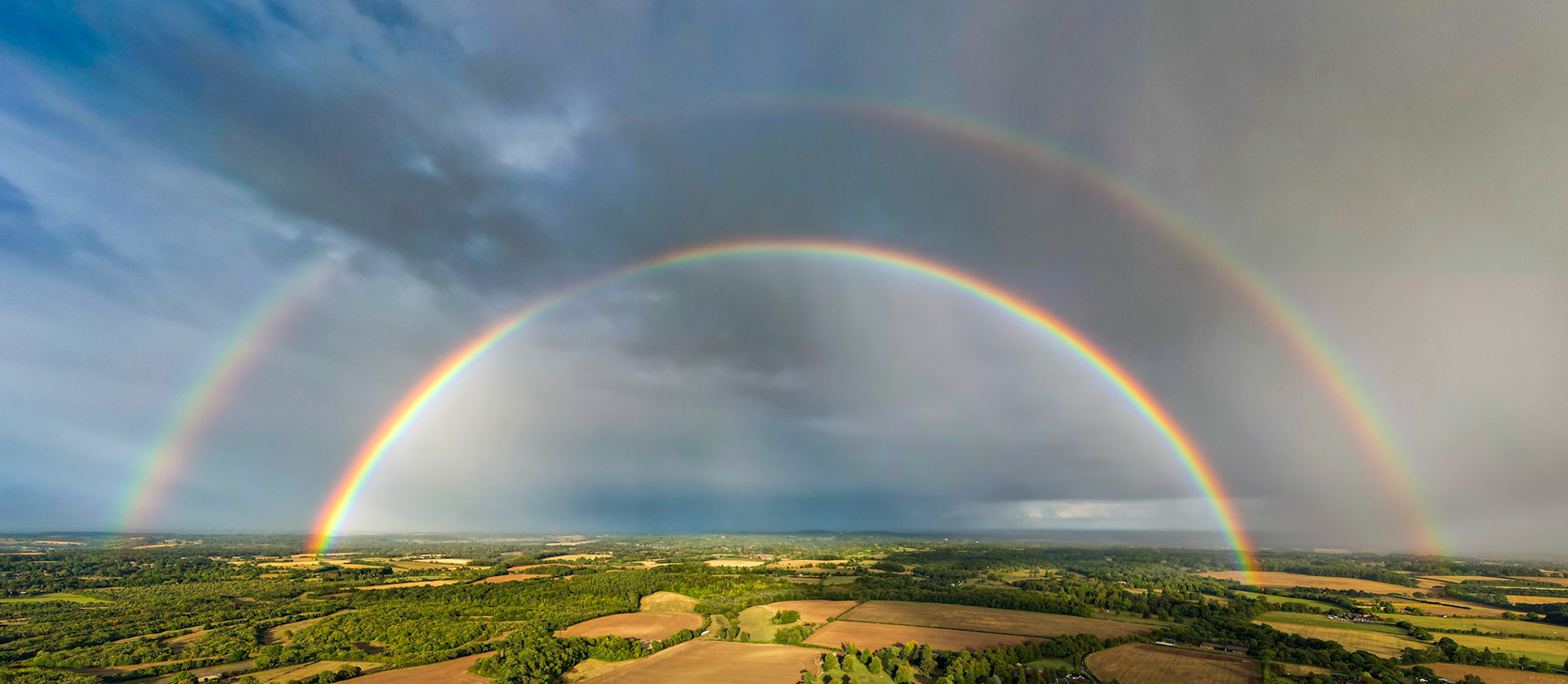A complete double rainbow spans the full width of the frame above the patchwork farmland and woodland of the Chilterns, photographed from altitude in the aftermath of a late-summer storm. Alexander's dark band is clearly visible between the primary and secondary bows, a rare degree of optical completeness seldom captured in a single unstitched aerial frame. The receding rain curtain to the right anchors the meteorological drama without sentimentality.