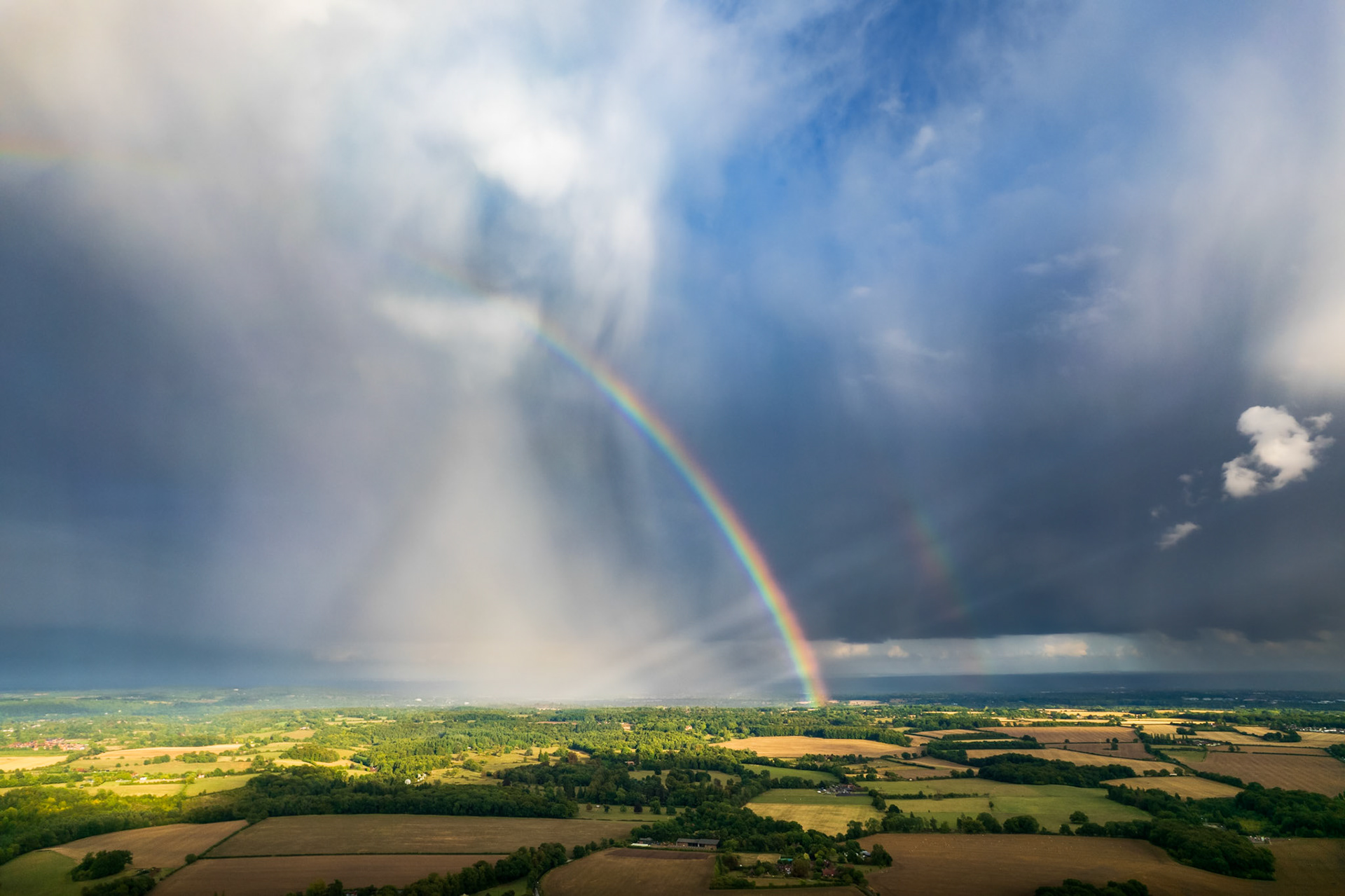 A vivid double rainbow descends through the base of a convective storm cell over the Thames Valley lowlands, captured from altitude as a rain shaft sweeps across a patchwork of summer fields and woodland. The aerial vantage reveals the full arc geometry impossible from ground level, set against a bruised, layered sky still broken by clear blue to the east. The proximity of sunlit crops and dark storm cloud gives the image an unusual tonal compression.