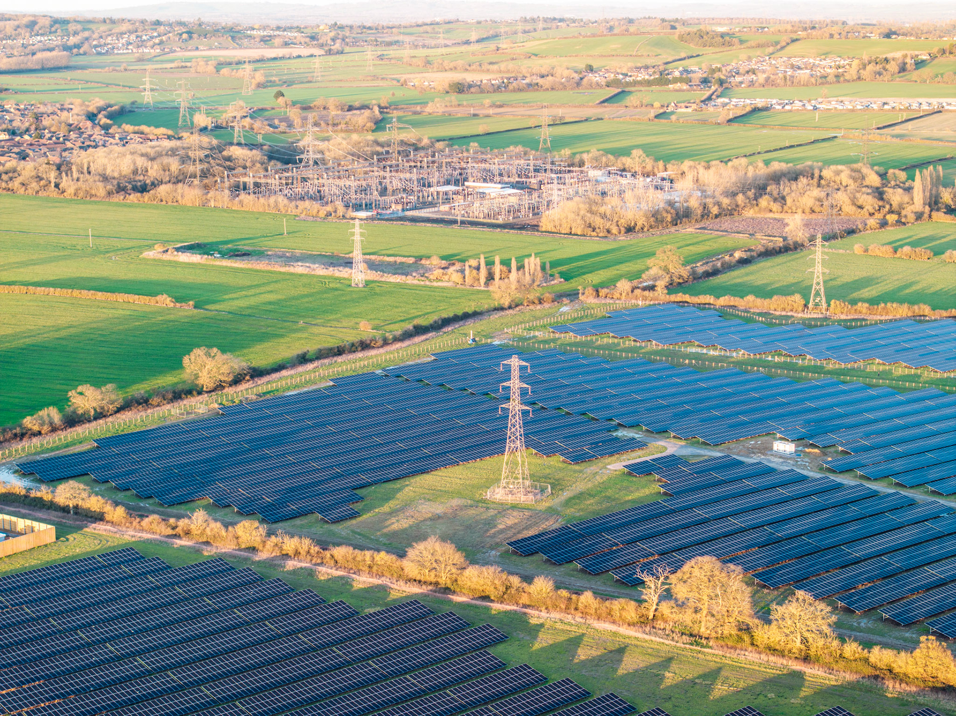 A large ground-mounted solar farm occupies former agricultural land on the outskirts of Oxford, Oxfordshire, its blue-black panel rows bisected by a transmission pylon connected to a visible grid substation beyond. Low winter sun rakes across the site from the south-west, picking out the geometry of the arrays against cropped grass margins and bare hedgerow trees. The composition records an energy-infrastructure landscape of growing familiarity in lowland England.