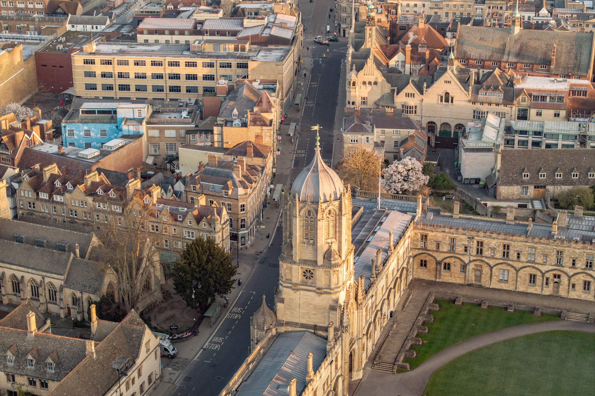 An aerial view across central Oxford catches the Tom Tower of Christ Church bathed in late-afternoon winter light, the pale Cotswold limestone glowing against the shadow-cooled roofscape of the High Street corridor. A single flowering cherry tree punctuates the collegiate stonework to the east. The composition is competent but the urban clutter beyond the college boundary dilutes the quiet authority the subject demands.