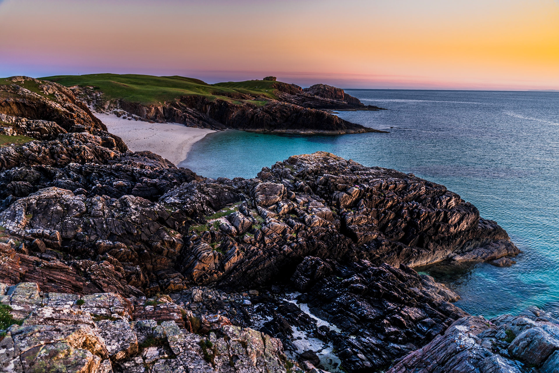 Sunset on Clachtoll beach, where the rugged coast meets the calm blue seas on the West coast of Scotland. The sky is warm in colour thanks to the setting sun.