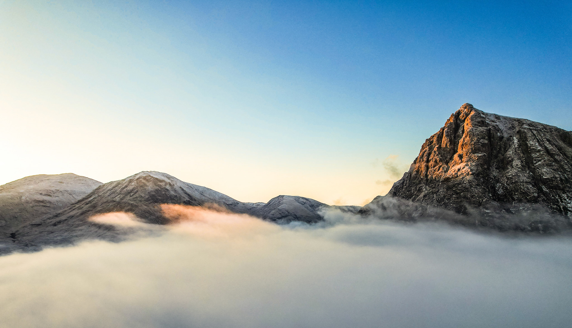 Snow-dusted summits break through a temperature inversion at first light, the valley floor buried beneath a still sea of cloud. Warm alpenglow catches the near rock face while the more distant peaks recede into pale silhouette. The aerial vantage compresses the layers into a clean, elemental arrangement of rock, cloud, and sky.