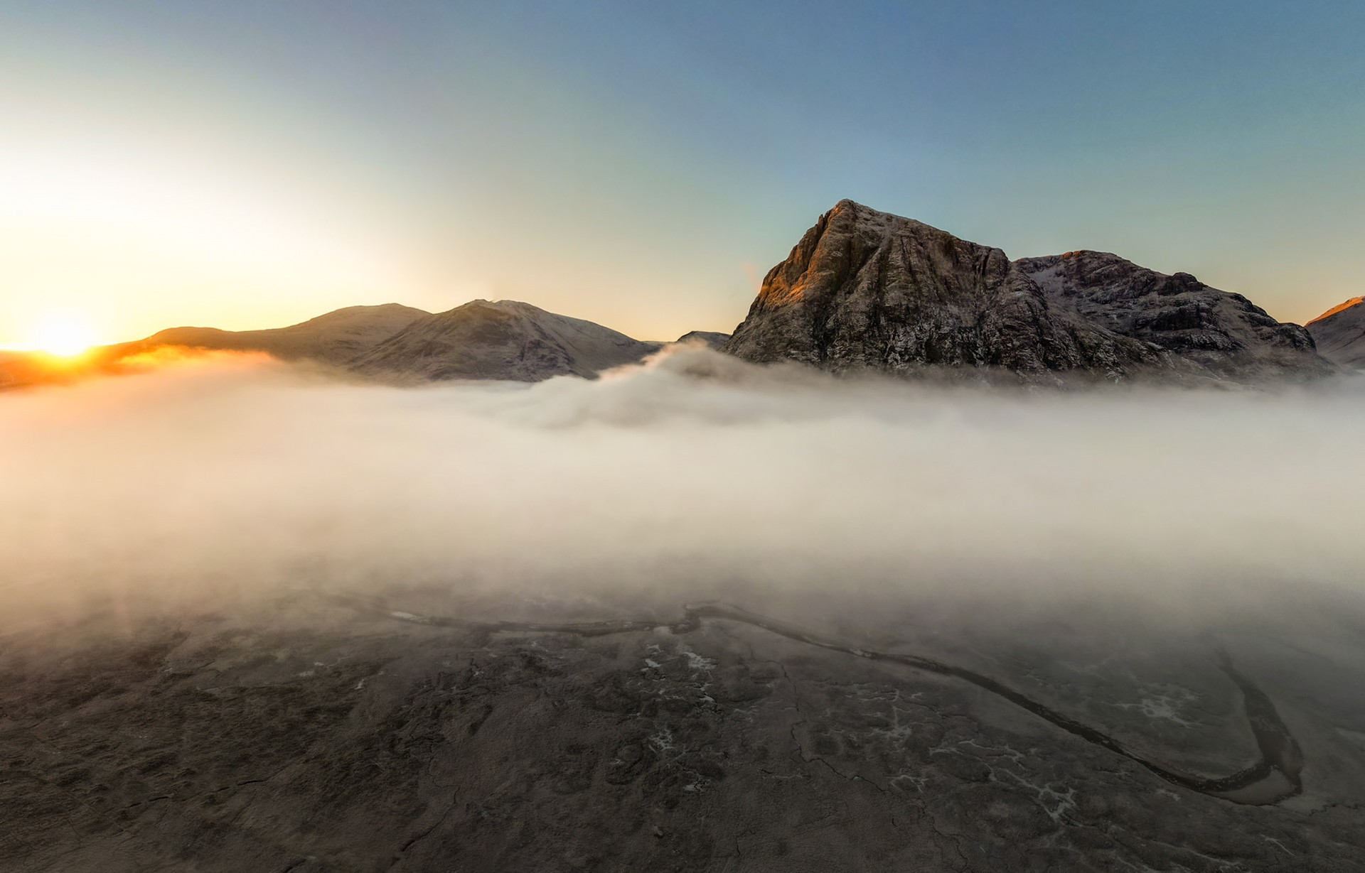 A temperature inversion fills the glen below Buachaille Etive Mòr at first light, the valley fog bisecting snow-dusted rock faces from a cold winter sky. Shot from altitude, the winding single-track road emerges from the mist base across frost-hardened moorland, anchoring the composition between the terrestrial and the atmospheric. The low sun grazes the peaks to the west, warming the summit ridges while the valley floor remains submerged.