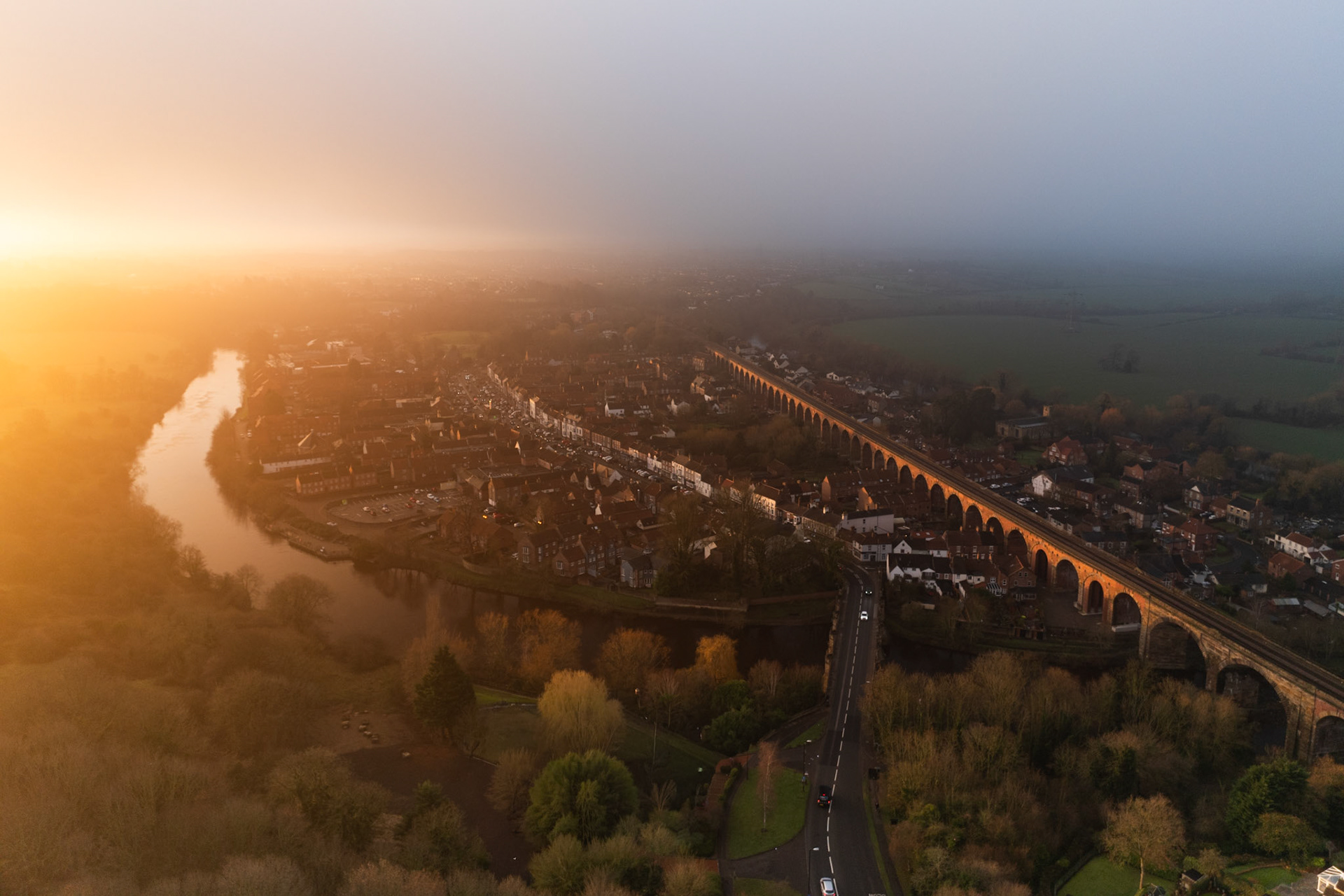 A Victorian railway viaduct bisects a river-bend market town as winter sunrise burns through low mist from the west, splitting the scene into warm gold and cool grey. The meander of the River Tees catches the light in a long reflection, anchoring the composition against the receding fog. Captured from altitude, the image holds a rare coincidence of architecture, weather, and light.