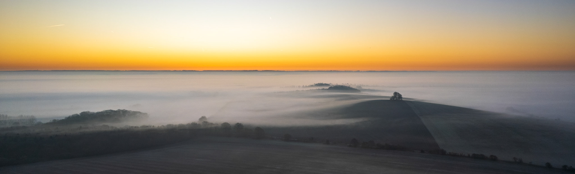 A temperature inversion fills the Vale of White Horse to the hilltops, leaving rounded chalk spurs as islands in a white sea. Photographed from altitude at first light near Abingdon-on-Thames on 1 March 2025, the pre-sunrise horizon holds a band of amber that grades cleanly into a cool blue sky, a crescent moon just visible above. The lone tree group on the nearest ridge provides the single point of scale and anchors an otherwise abstract composition.