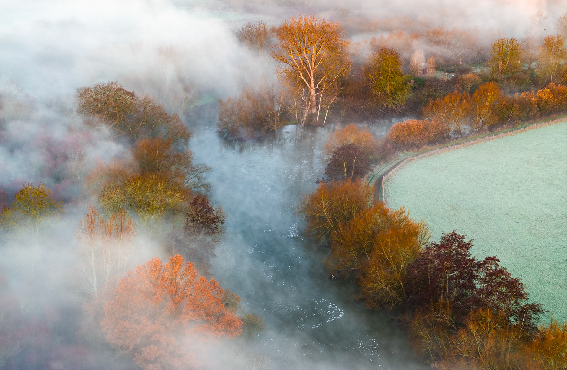 River mist pools between stands of late-autumn riparian woodland above the Thames near Abingdon, the vapour parting to reveal frost-silvered pasture on the valley edge. A single pale birch catches the first warmth of a December dawn, anchoring an otherwise dissolving scene. The aerial viewpoint compresses fog, foliage and frost into a composition of rare chromatic restraint.