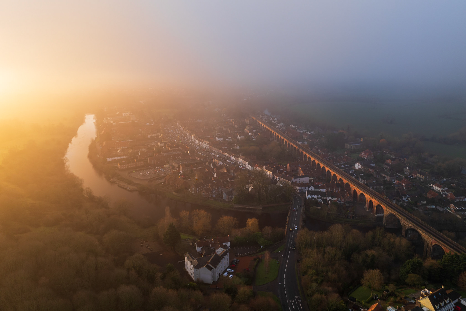 A low winter fog bisects the town, its edge drawn in a near-perfect diagonal across the rooftops and the long arched viaduct. Dawn light floods the western bank of the River Tees in amber while the eastern half of the frame remains suspended in cool grey. The thermal inversion catches a moment of accidental geometry between warmth and cold.