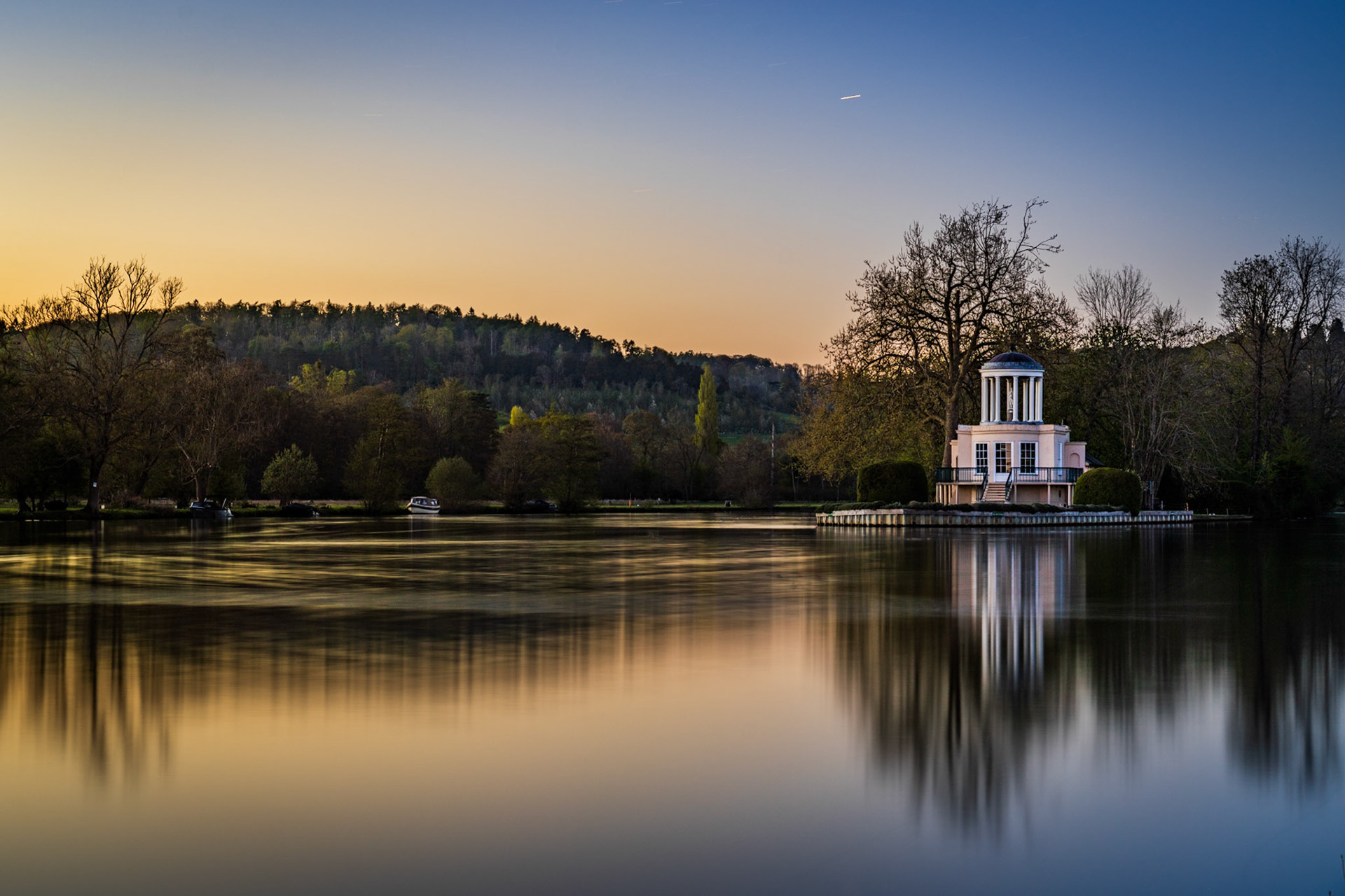 A neoclassical riverside temple catches the last warmth of an April sunset on the Thames near Henley, its columns and dome reflected in long-exposure-smoothed water. The gradient from amber horizon to deep blue zenith frames the building with quiet formality. A vapour trail and moored boats add understated human presence without disrupting the stillness.