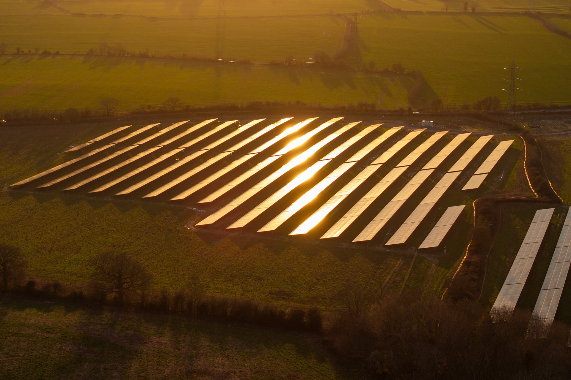 Rows of photovoltaic panels catch the last angle of winter sun near Oxford, their glass surfaces flaring into a concentrated column of reflected light. Seen from altitude, the geometry of the installation resolves into something closer to abstraction than infrastructure. The surrounding fields and hedgerows anchor the composition in the English agricultural lowlands.
