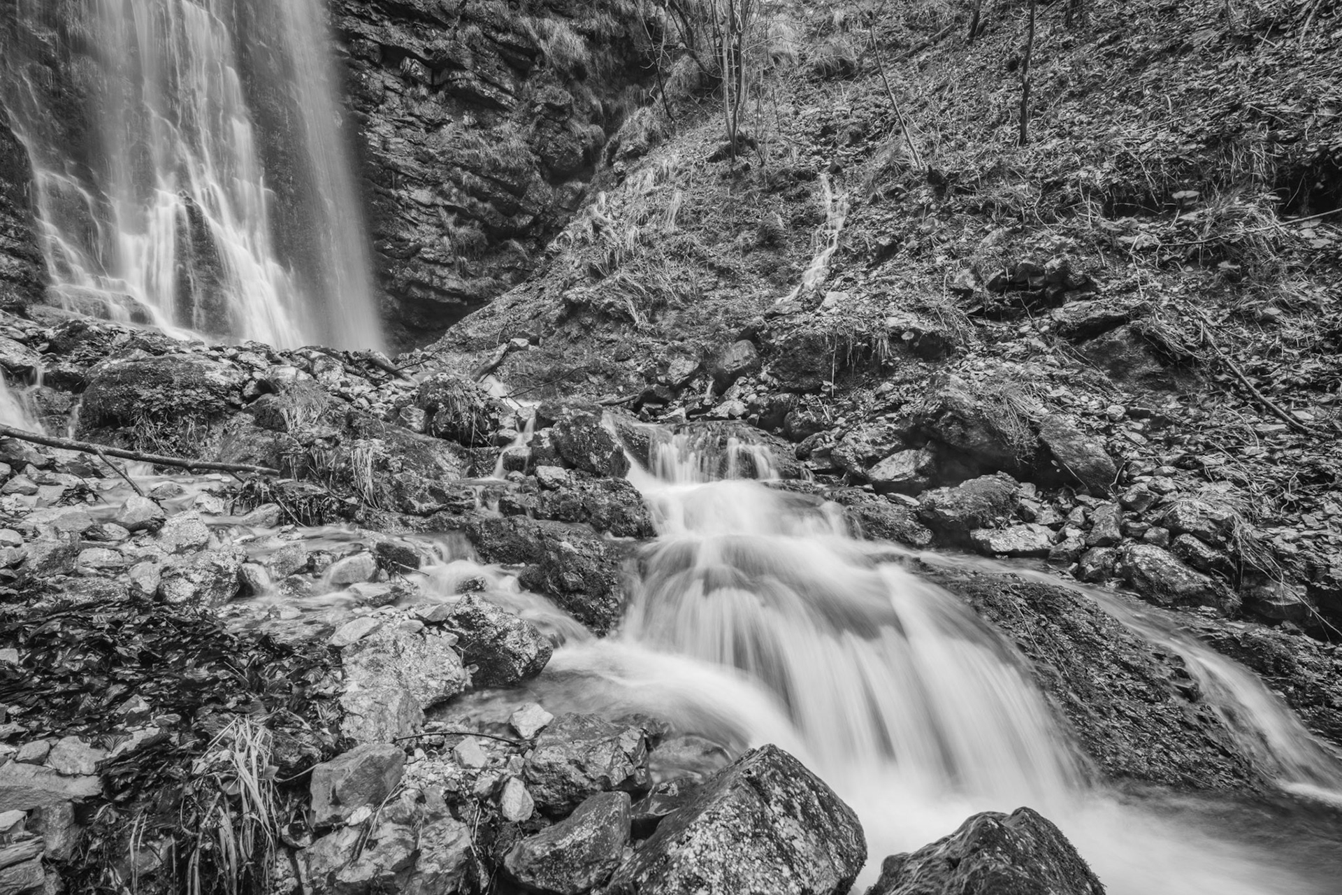 A long exposure compresses the energy of a hillside waterfall into soft tonal bands, the silked water contrasting with the coarse texture of wet rock and loose scree below. The image works the foreground cascade well but the mid-ground is busy and lacks a clear resting point for the eye.