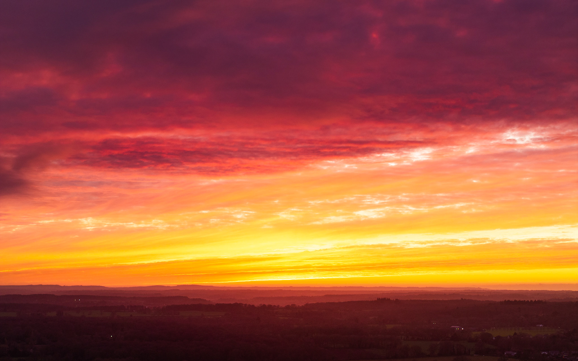 A drone vantage above the Thames Valley records the final seconds of dawn colour before winter sunrise, the sky grading from deep crimson through amber to a luminous yellow band at the horizon. Layers of low cloud catch the light in horizontal strata while the darkened valley floor below retains scattered settlement lights. The composition is dominated by sky, leaving the landscape as a minimal anchor.